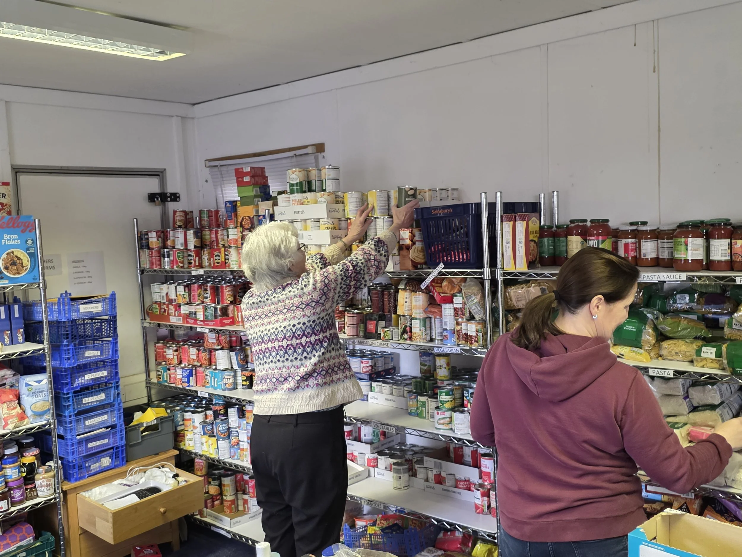 Two women shopping for canned and jarred goods on store shelves, one reaching for a can, the other examining products.