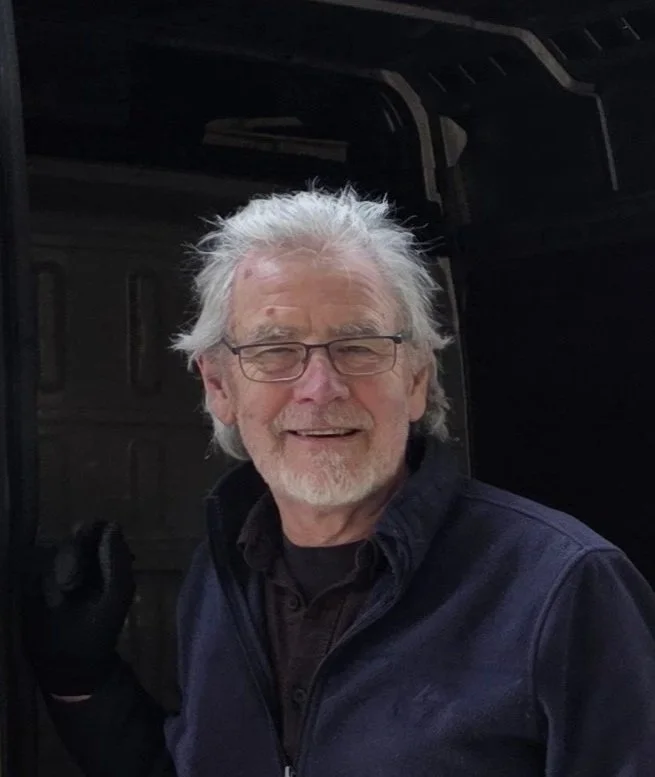 An elderly man with white hair, glasses, and a beard smiling and wearing a black jacket, standing next to a black vehicle.
