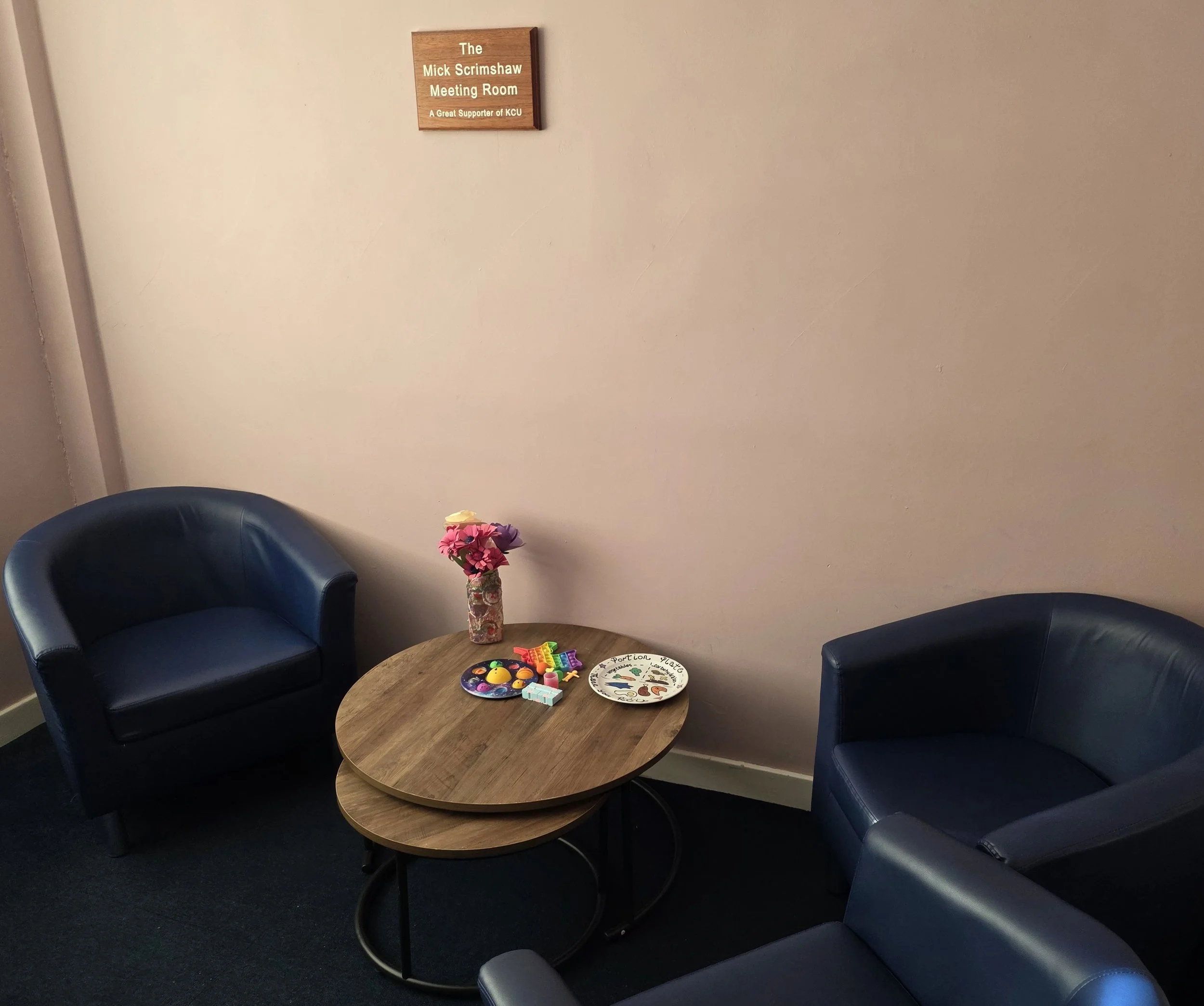Waiting area with two dark blue leather armchairs, a wooden table with a vase of pink flowers, a colorful toy set, and a decorative plate, against a plain beige wall with a wooden sign that reads 'The Mick Scrimshaw Meeting Room'