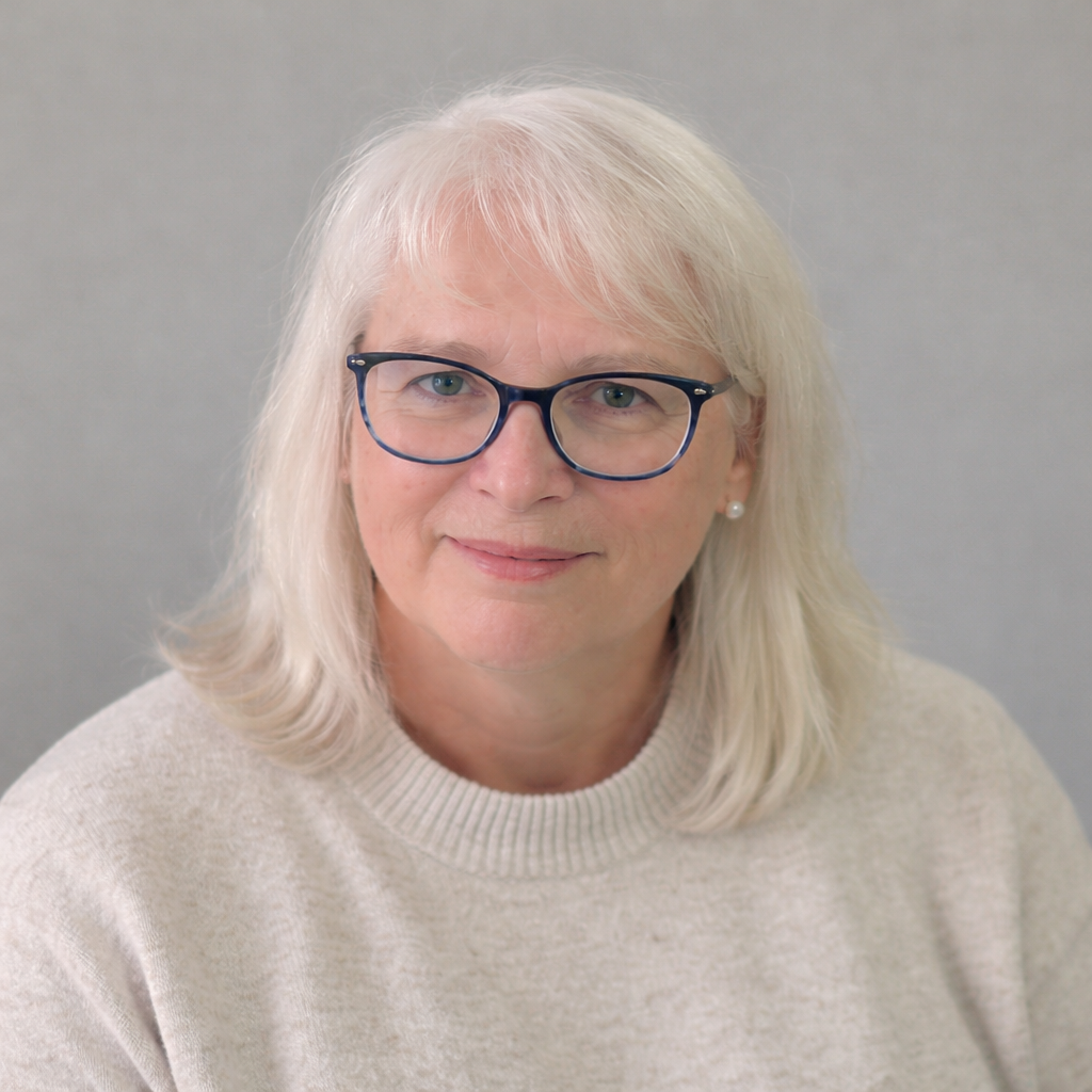 A portrait of a woman with blonde hair, wearing blue glasses, a pearl earring, and a beige sweater, smiling softly against a plain gray background.