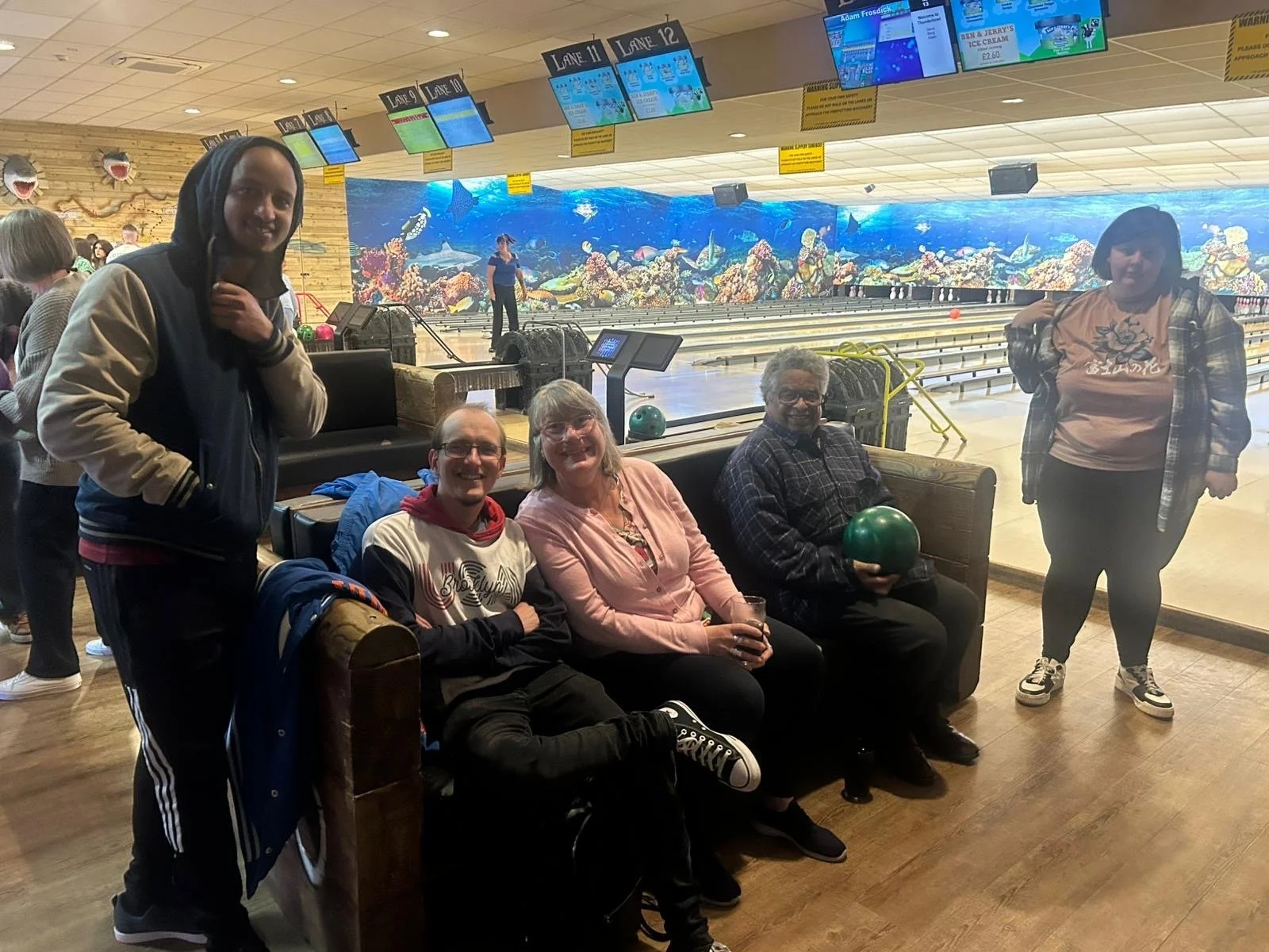Five people at a bowling alley, sitting and standing in front of bowling lanes. Two women standing on either side, three people sitting on a bench, one holding a bowling ball, another holding a drink. Background includes a large underwater-themed mural and electronic scoreboard displays.