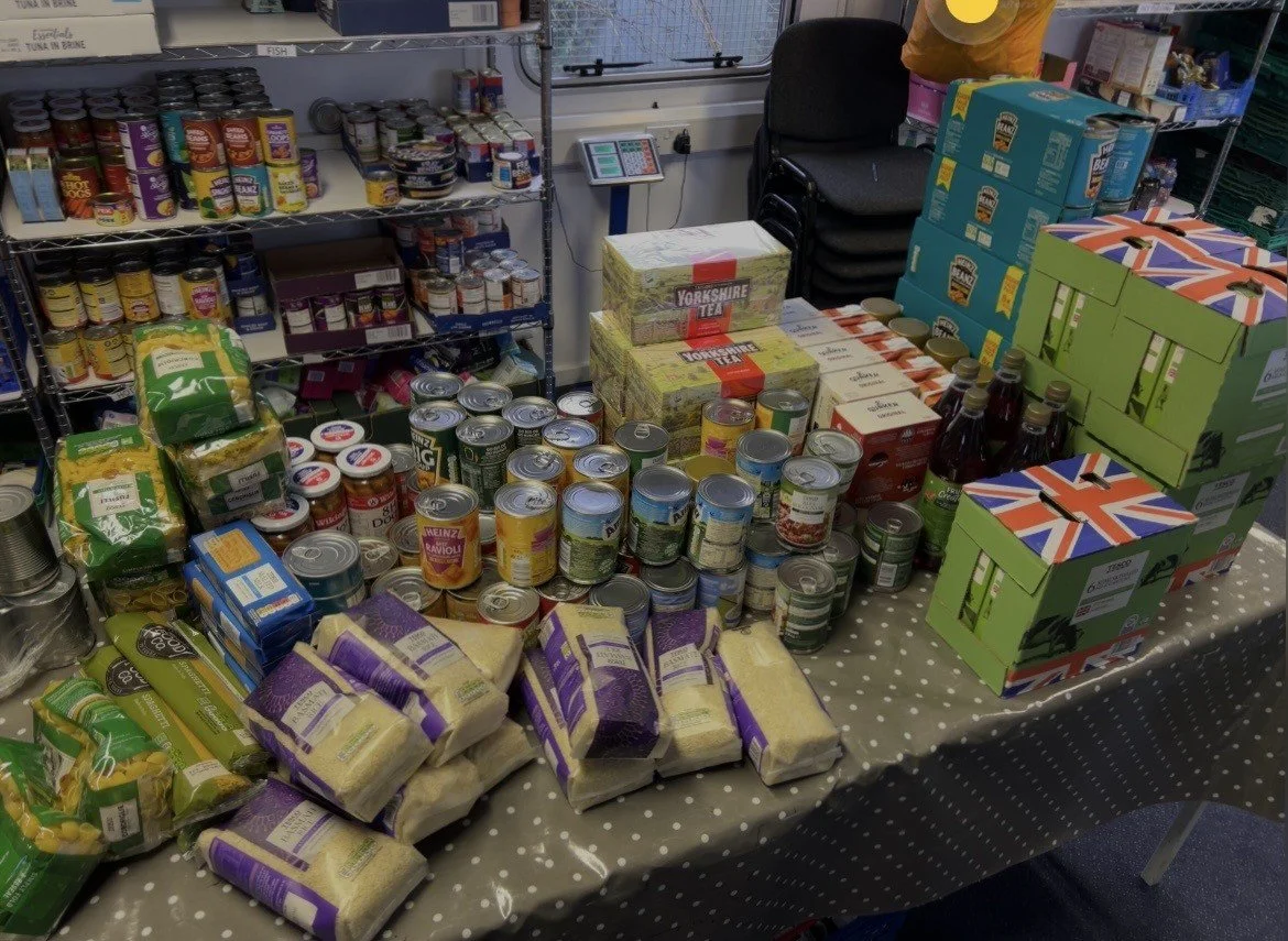 Table of canned and packaged food items for a food drive or donation event, with boxes of tea, bags of pasta, cans of soup, and bottled beverages.