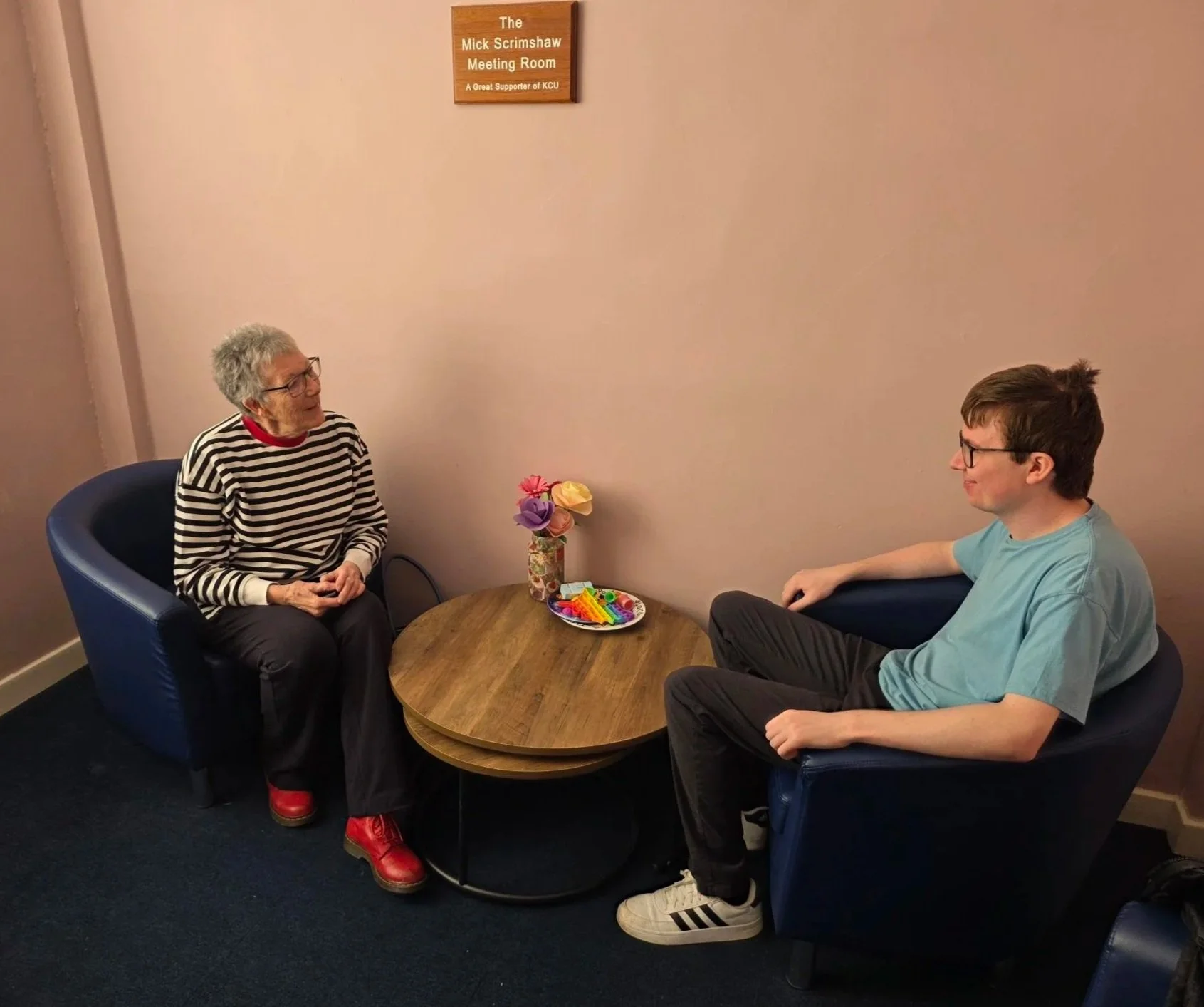 An elderly woman and a young man sit facing each other in a small meeting room with a sign on the wall that reads 'The Mick Scrimshaw Meeting Room.' The woman has gray hair, glasses, and is wearing a striped shirt, black pants, and red shoes. The young man has brown hair, glasses, and is wearing a light blue t-shirt, dark pants, and white Adidas sneakers. A wooden table with a vase of colorful paper flowers and a plate of colorful candies is between them.