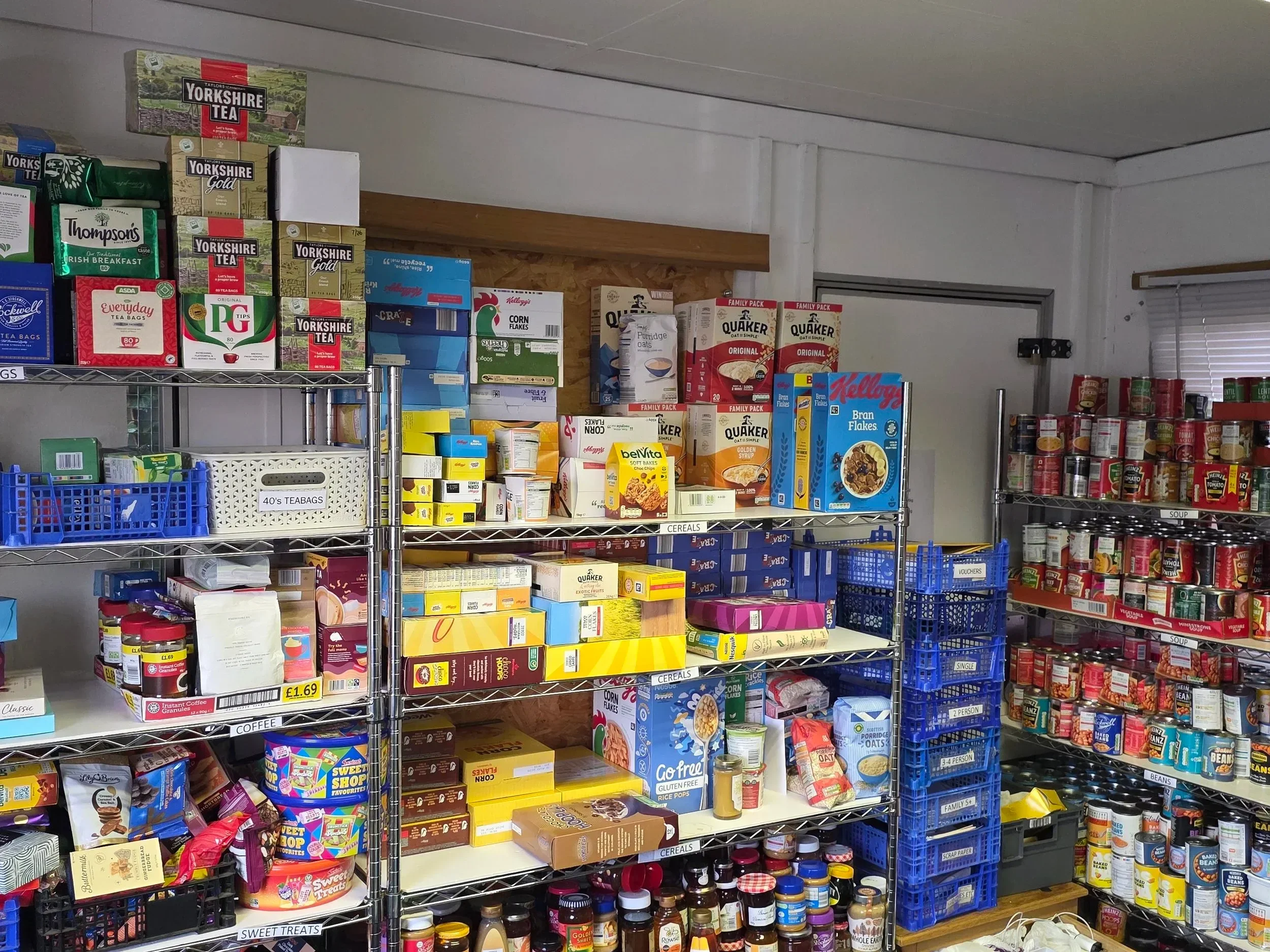 Shelves stocked with various canned foods, boxes of cereal, tea, coffee, and pantry items in a grocery store aisle.