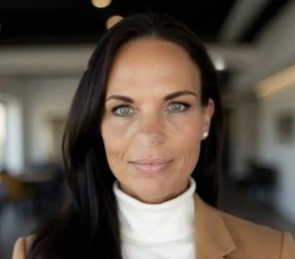 A woman with dark hair and blue eyes wearing a white turtleneck and beige blazer, indoors with blurred background.
