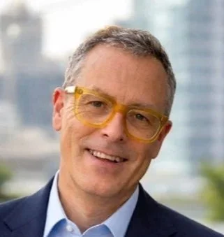 Portrait of a smiling man with yellow glasses, wearing a suit, with a cityscape background.
