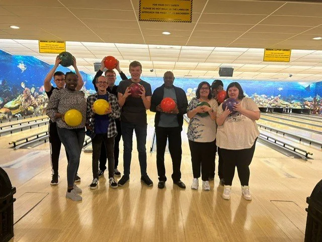 Group of eight people at a bowling alley, holding colorful bowling balls and posing for a photo.