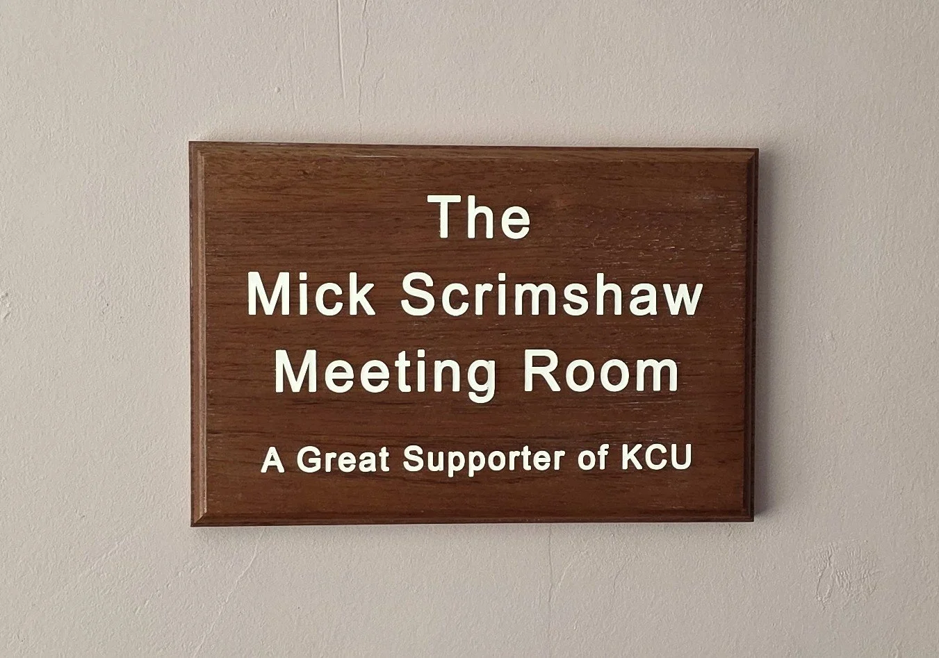 Wooden sign on wall reading 'The Mick Scrimshaw Meeting Room - A Great Supporter of KCU'.