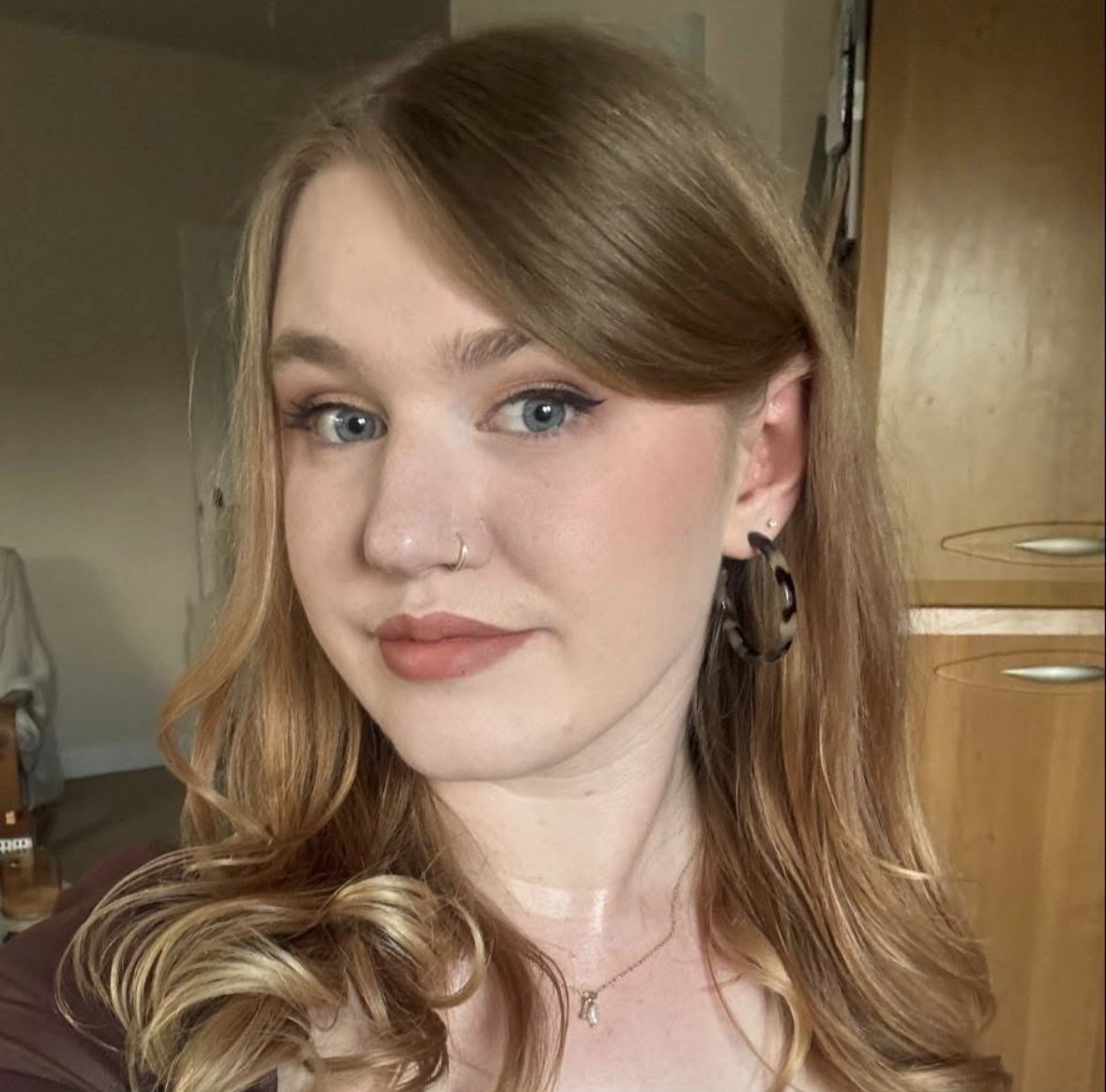 Close-up portrait of a young woman with reddish-brown hair, blue eyes, and light makeup, wearing a black earring and a small necklace, indoors with wooden furniture in the background.