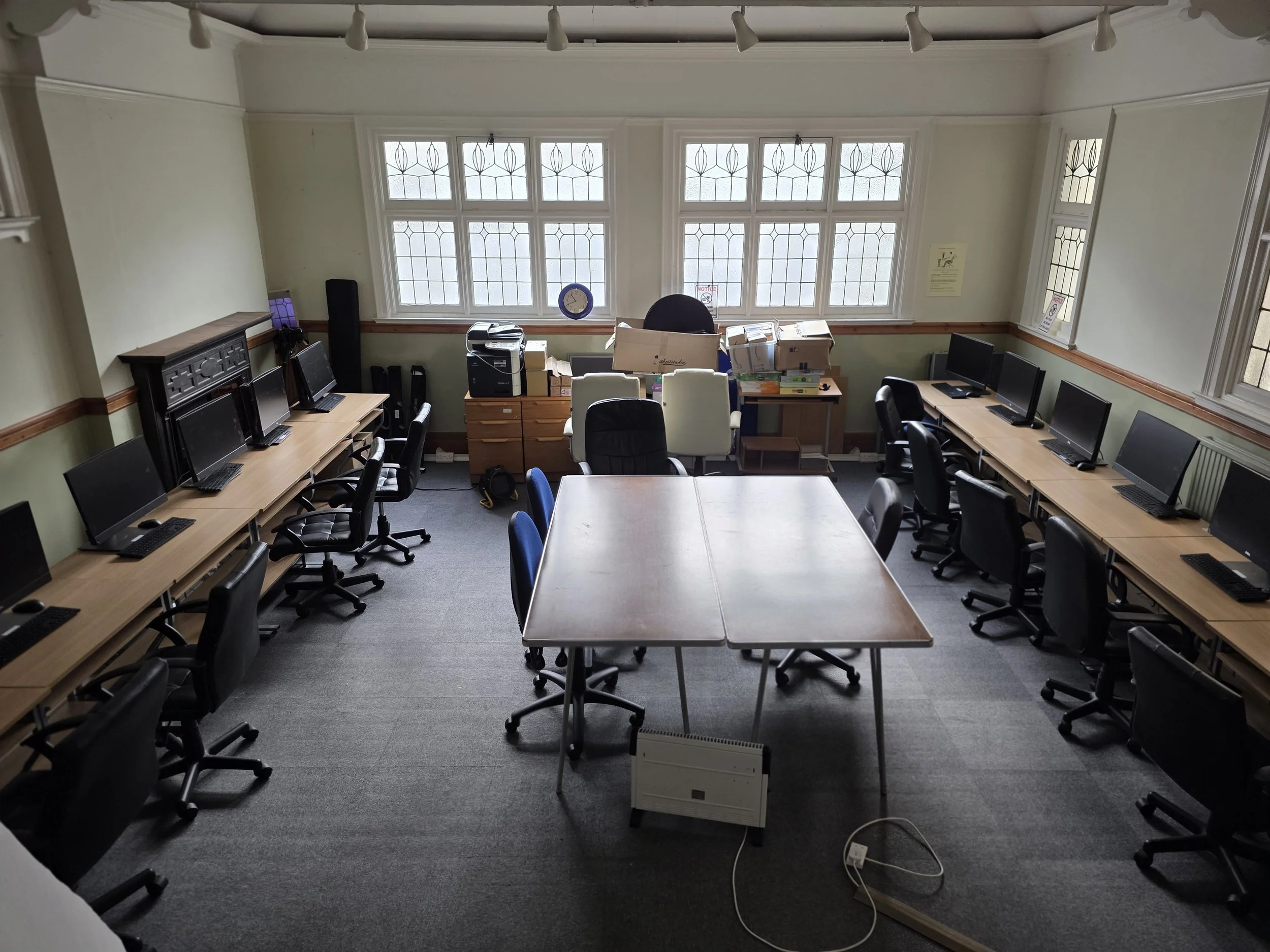 An empty computer training room with 8 desktop computers on long desks arranged along the walls and 4 chairs at each desk. A large table is in the center of the room. There is a window wall with frosted glass and a clock on the window ledge. The room has a light green wall color with wooden wainscoting and gray carpeted floor.