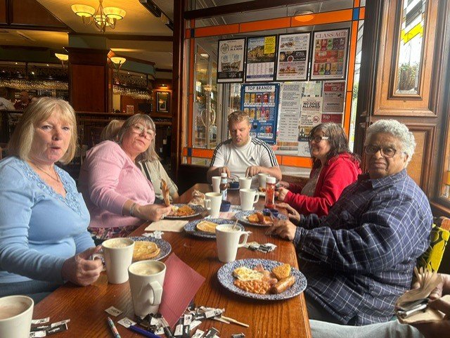 Five people sitting at a wooden table in a restaurant, eating breakfast with plates of eggs, toast, and pastries, and coffee mugs, with a window and posted notices in the background.