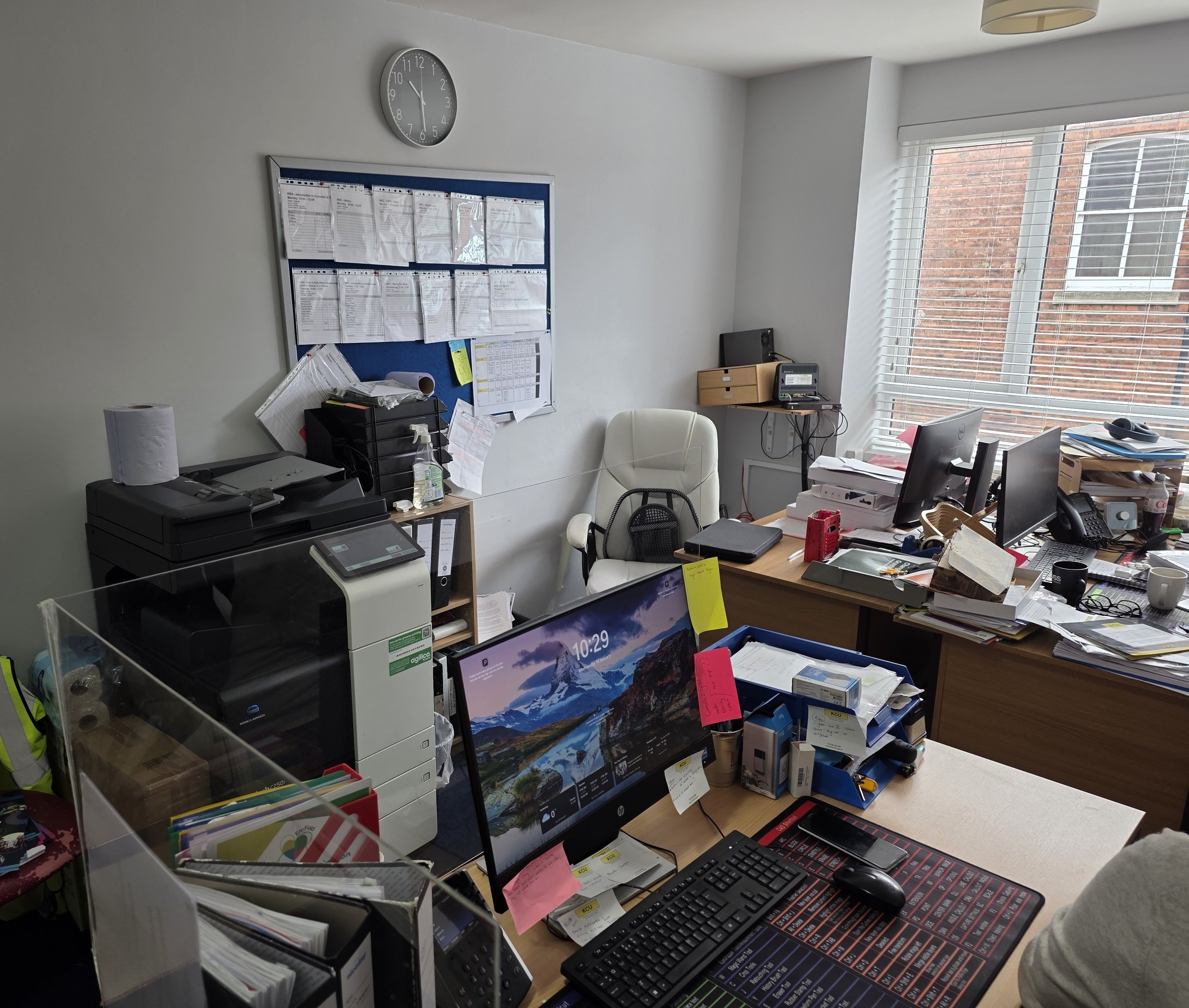 Office environment with cluttered desk, multiple monitors, printer, and papers, with a window showing a brick building outside.