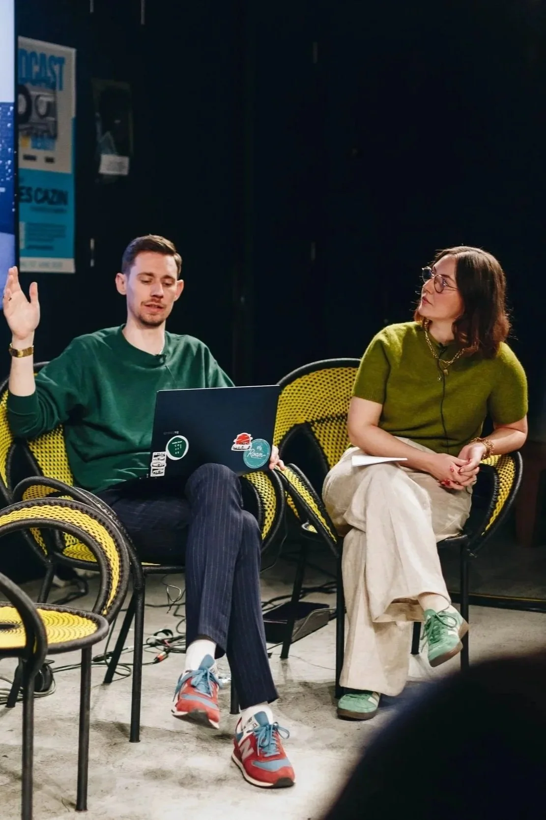 Two people sitting in yellow and black chairs, having a discussion. The man on the left, with a laptop on his lap, appears to be speaking with his hand raised. The woman on the right is listening, holding a notebook, wearing glasses, and dressed in an olive green top and beige pants.
