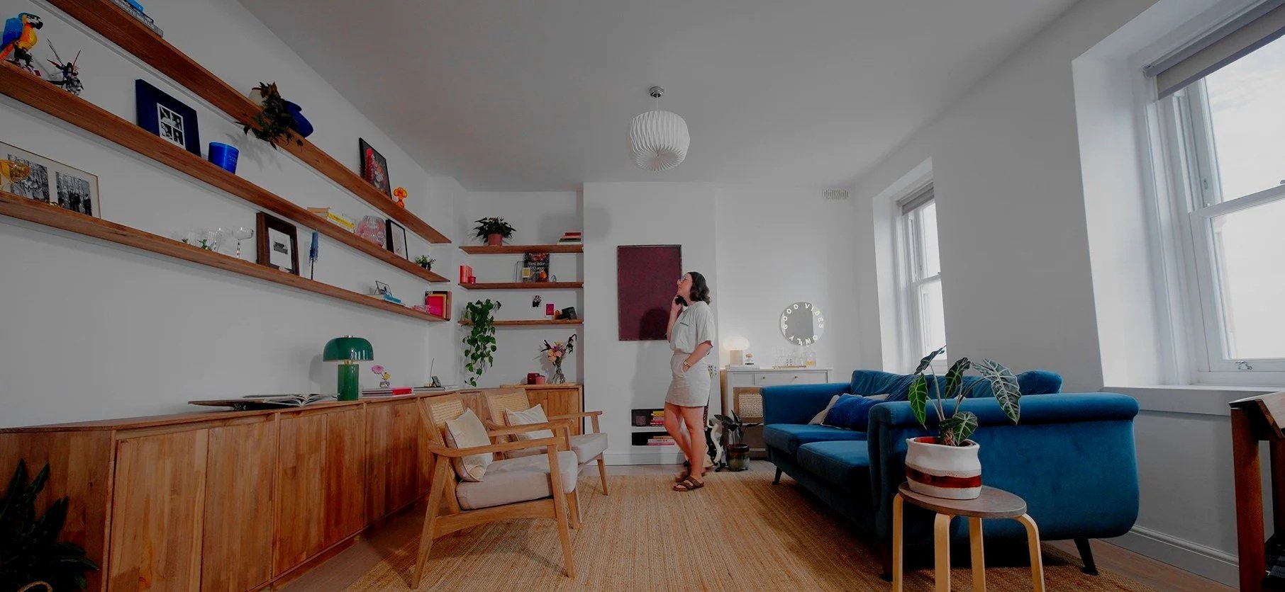 A woman standing in a bright living room, talking on a phone. The room features a blue sofa, wooden chairs with cushions, a wall with floating shelves holding decorative items and books, and large windows letting in natural light.