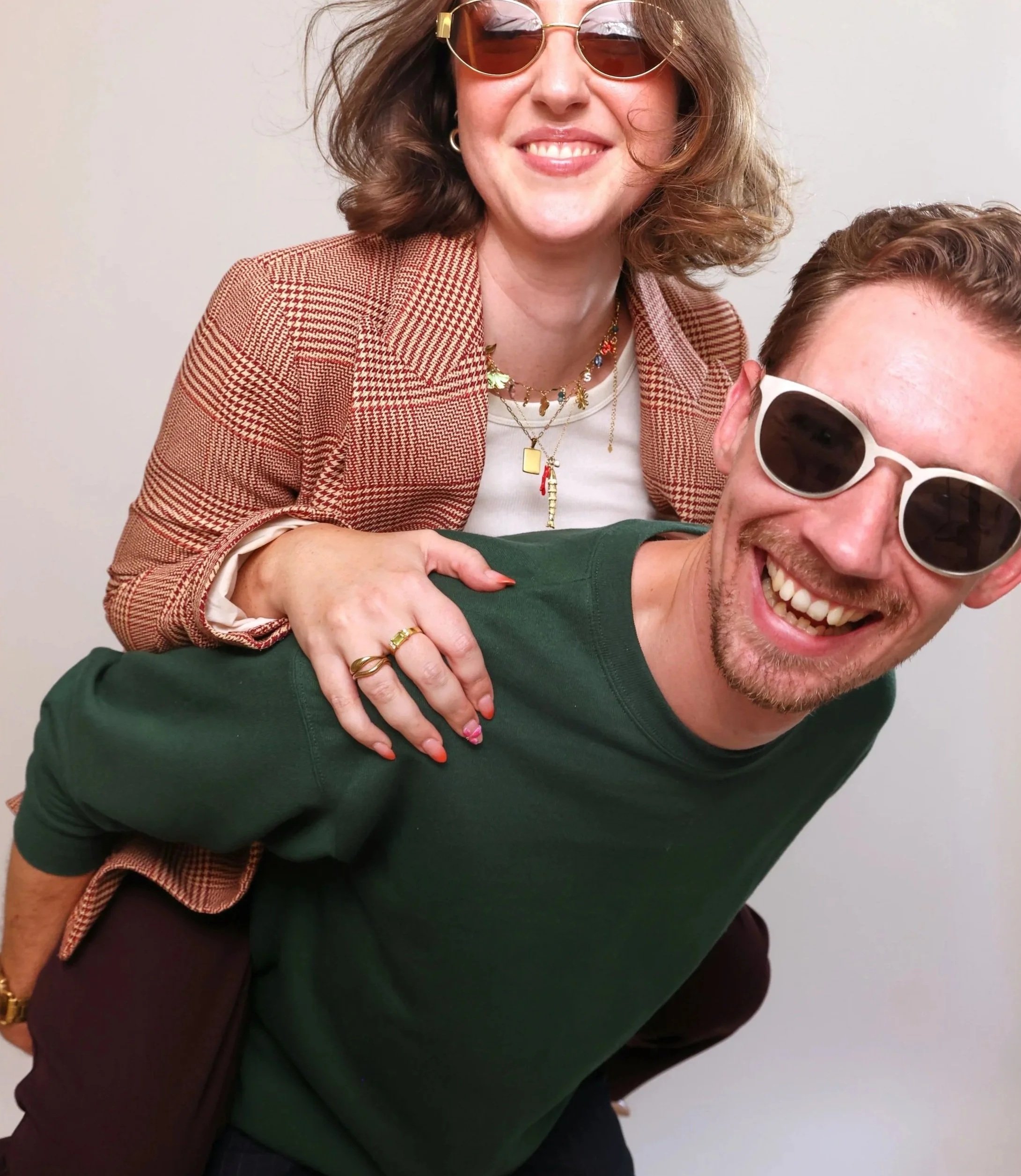 A woman with curly hair, wearing sunglasses and a checkered blazer, is smiling and leaning on a man. The man, with short hair, a goatee, and sunglasses, is also smiling widely. They are posing against a plain background.