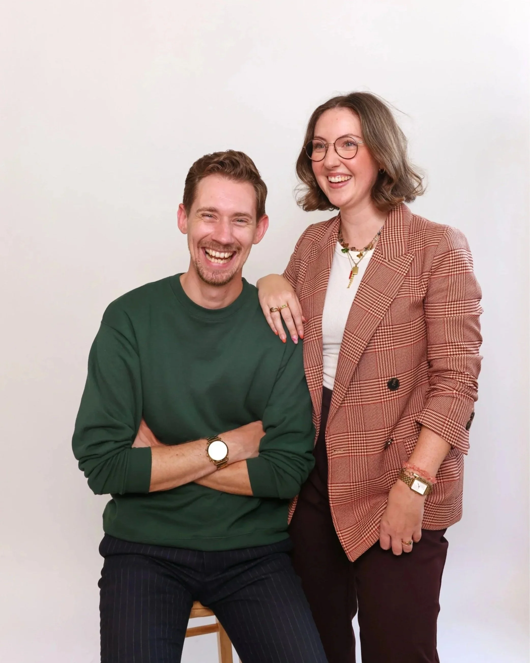A man and woman smiling. The man is sitting on a stool wearing a green sweatshirt and black pants. The woman is standing beside him wearing a checked blazer, black pants, and glasses, with her hand on his shoulder.