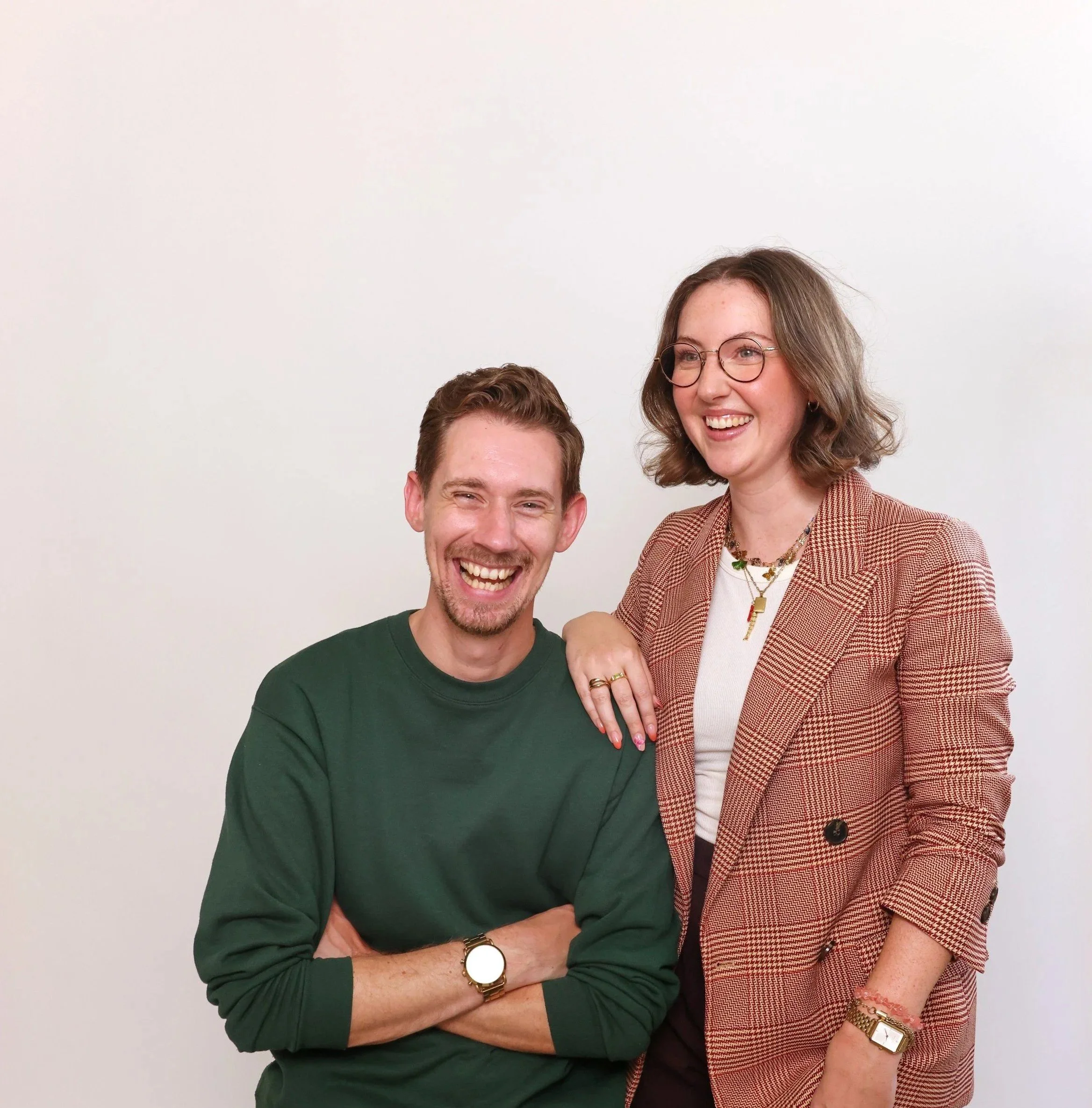 A man and woman smiling and posing together against a plain white wall. The woman has her hand on the man's shoulder.