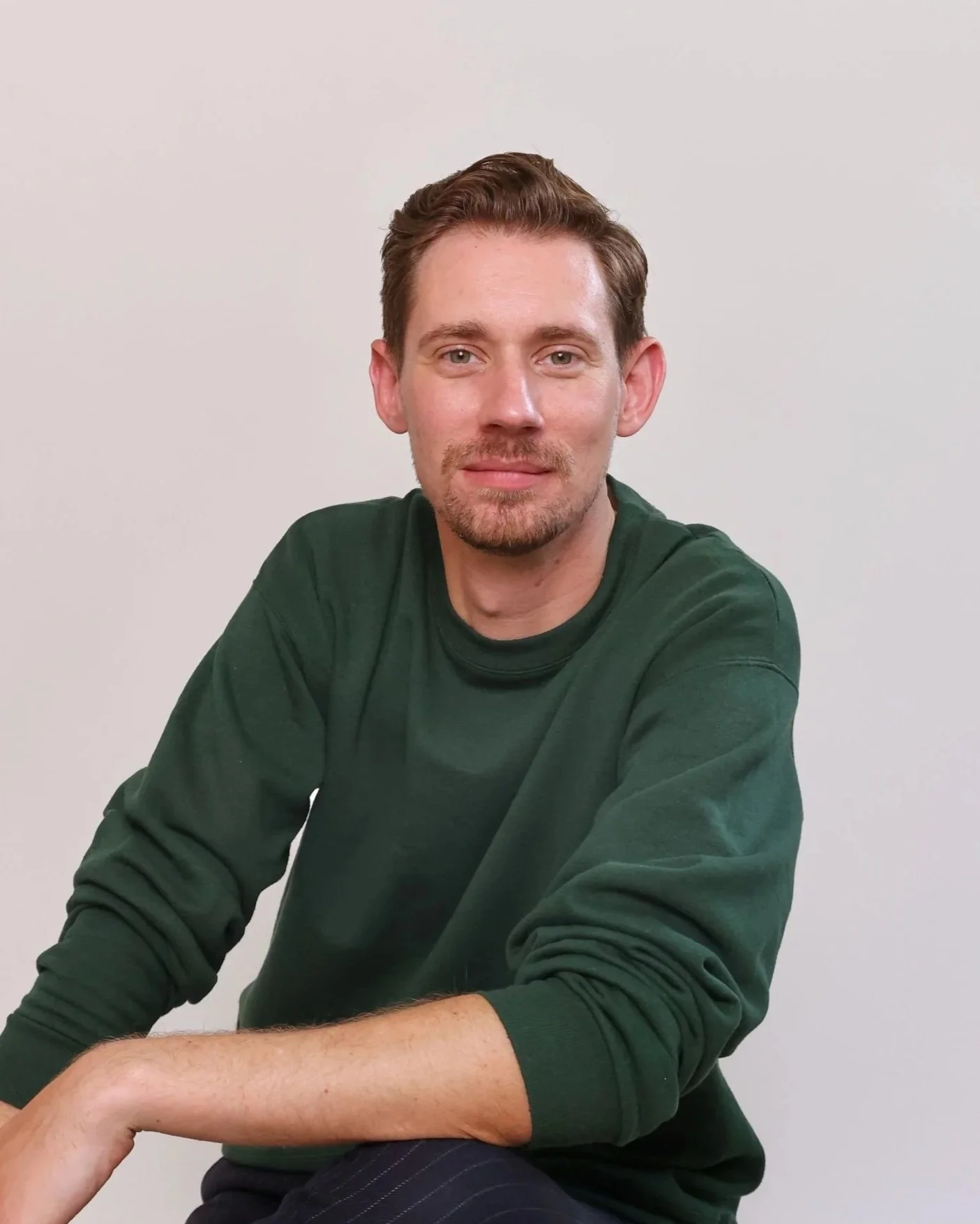 A man with short brown hair, a light beard, and wearing a dark green sweater, sitting against a plain white background.