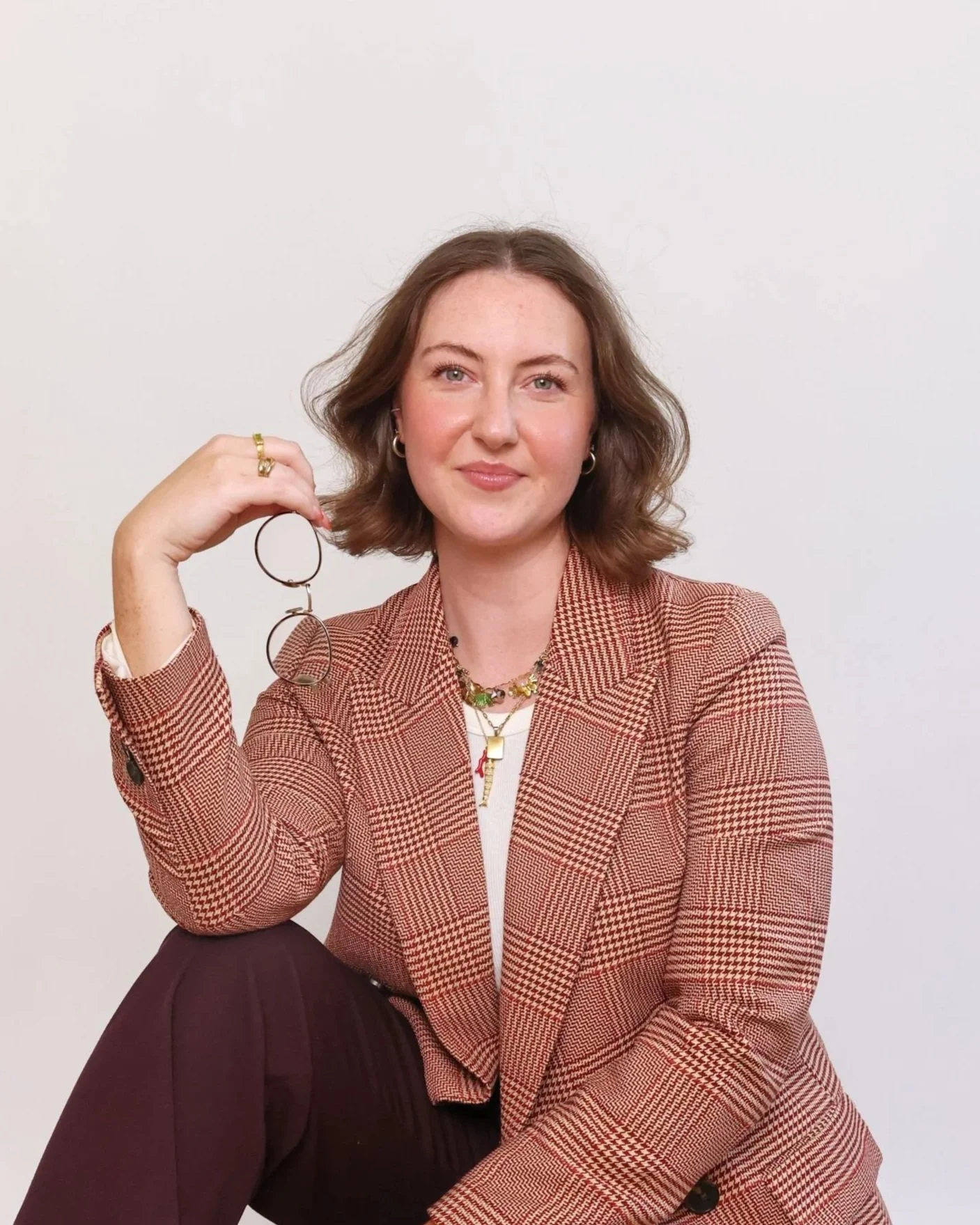 A woman with shoulder-length brown hair smiling and holding glasses, sitting against a plain white background, wearing a patterned blazer and colorful jewelry.
