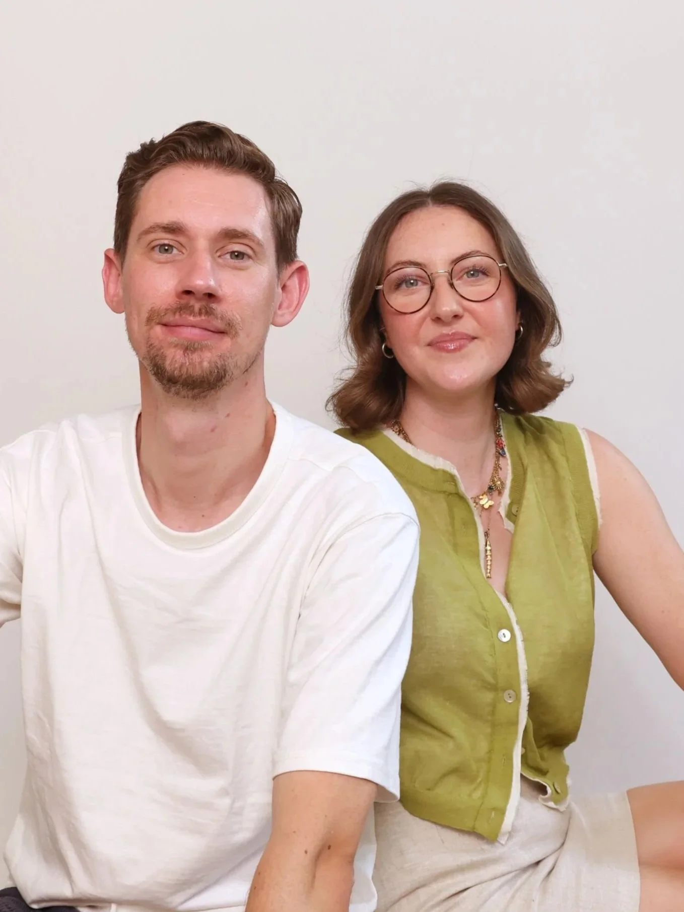 A man and woman sitting together against a white wall, looking at the camera. The man has brown hair, a beard, and is wearing a white t-shirt. The woman has shoulder-length brown hair, glasses, and is wearing a sleeveless green vest and beige shorts.