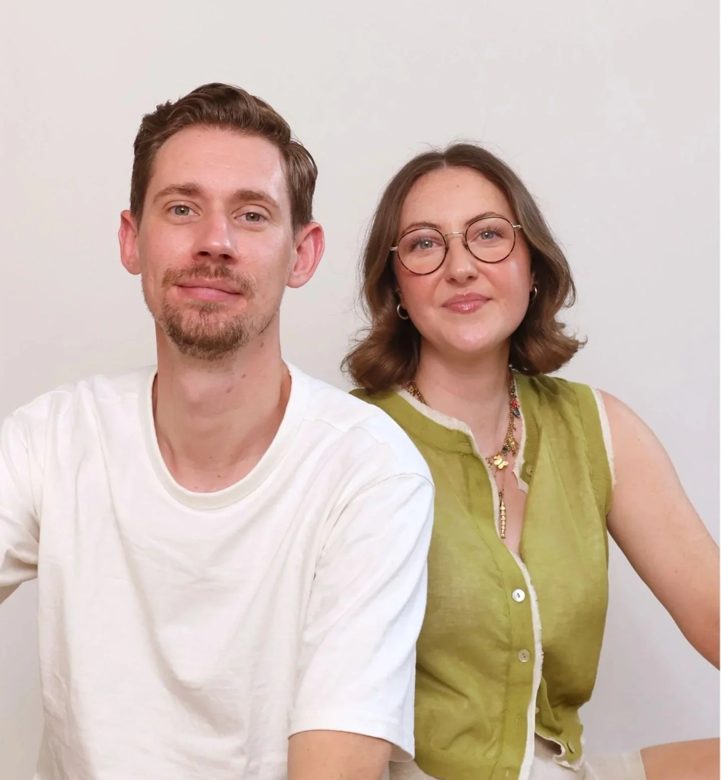 A young man with short brown hair and a goatee, wearing a white t-shirt, sitting next to a young woman with shoulder-length brown hair, glasses, and wearing a sleeveless green shirt, both smiling against a plain white background.
