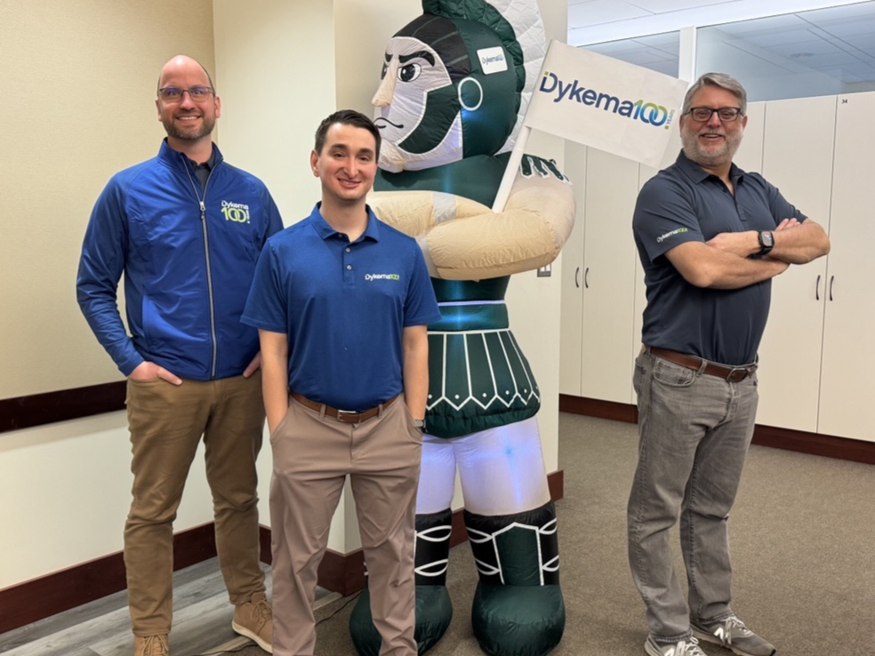 Group of Dykema staff posing with the Spartan Mascot in the firm's Lansing office wearing their Dykema100 swag to celebrate the firm's centennial. 