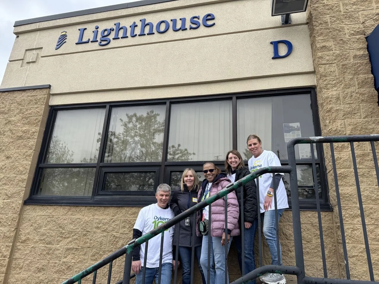 Group of five people standing on a staircase in front of a building with a sign that reads 'Lighthouse'. Four are women and one is a man, all smiling and wearing casual clothing, some in T-shirts with 'Dyke' written on them.