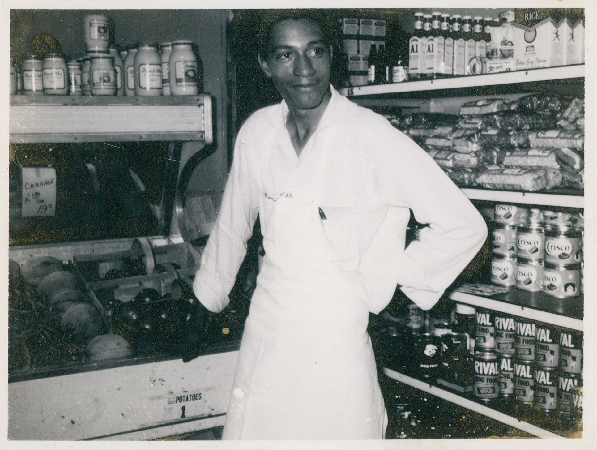 A man in a white apron standing in a grocery store aisle with shelves of canned goods, rice, and canned vegetables in the background.