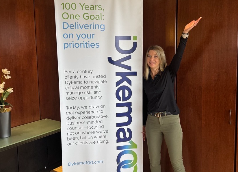A woman standing next to a tall vertical banner that reads '100 Years, One Goal: Delivering on your priorities.' The woman has blonde hair, is smiling, and raising her right arm. She is wearing a black shirt and khaki pants. The background features a