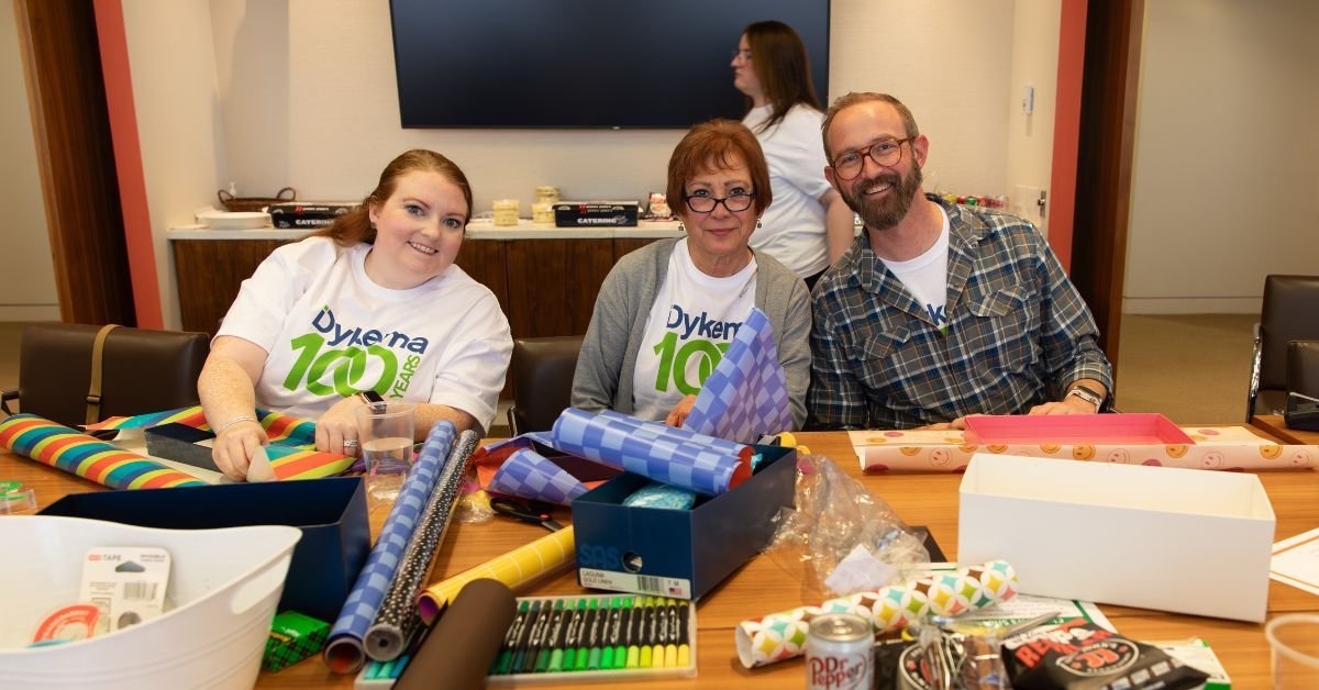 Three people sitting at a table working on wrapping gifts with wrapping paper and supplies, celebrating a 100th anniversary event for Dykemar.