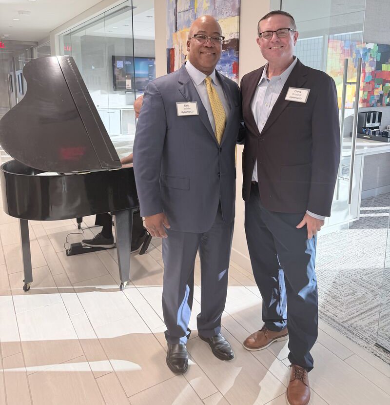 Two men in suits smiling and standing in front of a room with a black grand piano and colorful abstract artwork on the wall.