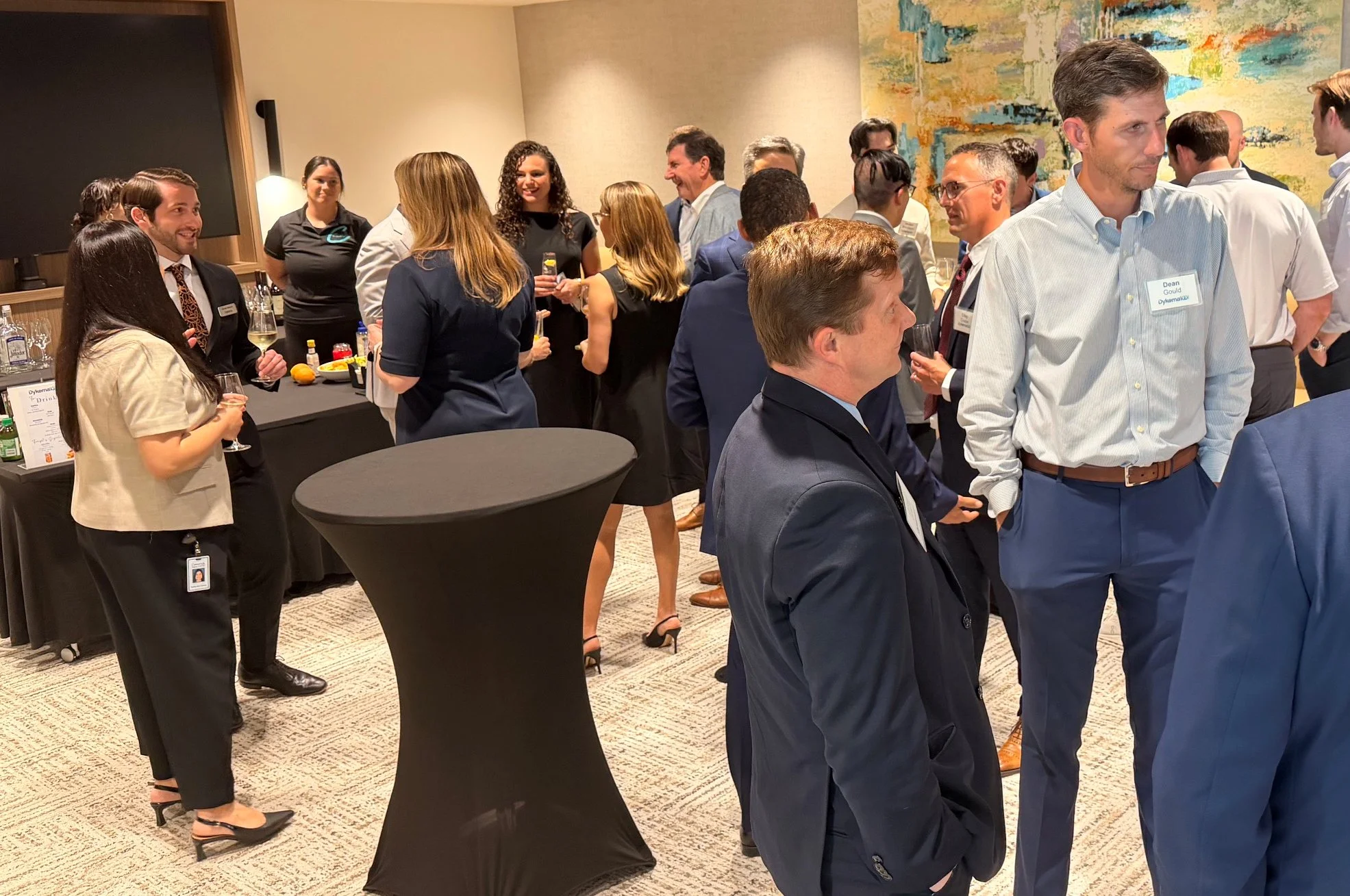 Business professionals at a networking event, engaging in conversation, with some holding drinks, in a room with colorful abstract art on the wall, and high-top tables.