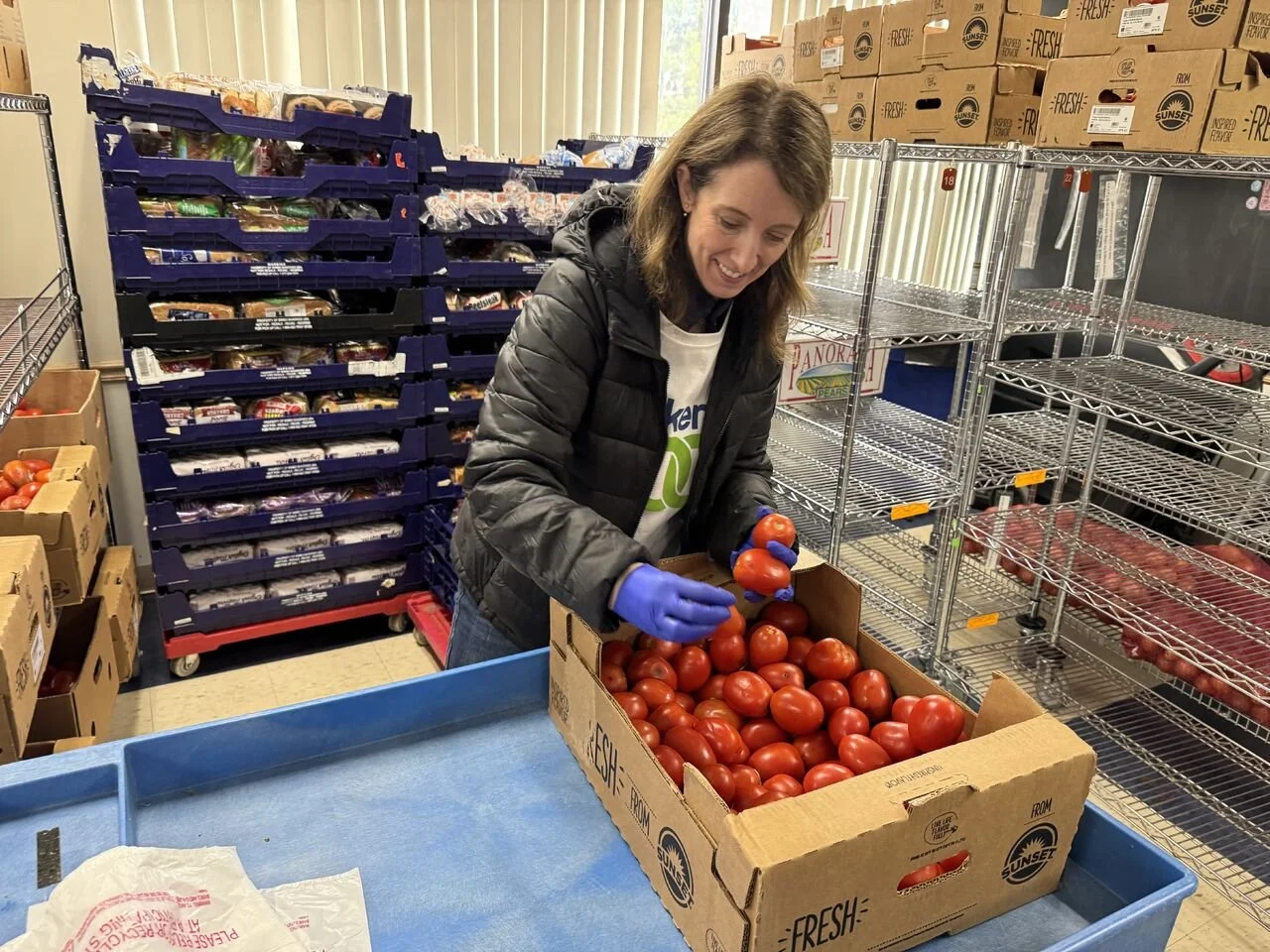 A woman packing fresh tomatoes into a cardboard box in a grocery store or warehouse.