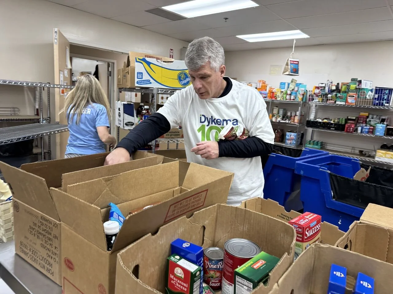 A man wearing a white T-shirt with the text 'Dykema 10' is organizing food items into cardboard boxes on a table in a food bank. A woman in a blue shirt is in the background, also working with food on shelves.