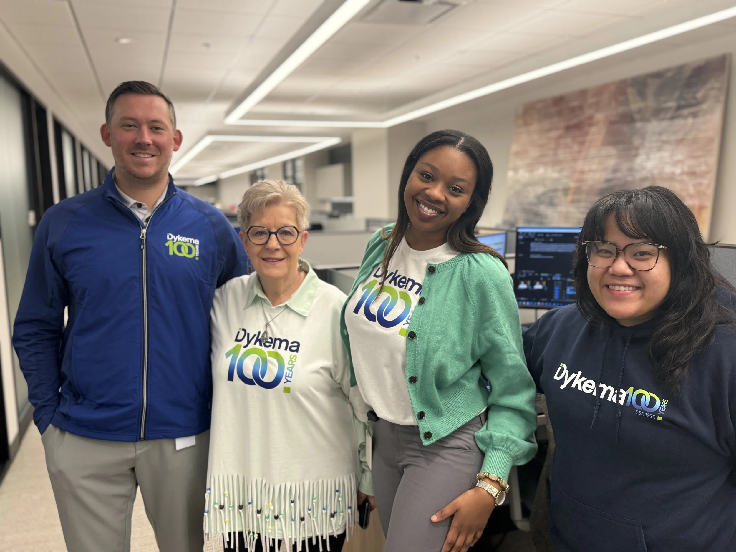 Group of four Dykema staff smiling in the firm's Detroit office, wearing shirts and hoodies with a 'Dykema 100 Years' logo, celebrating the company's centennial.