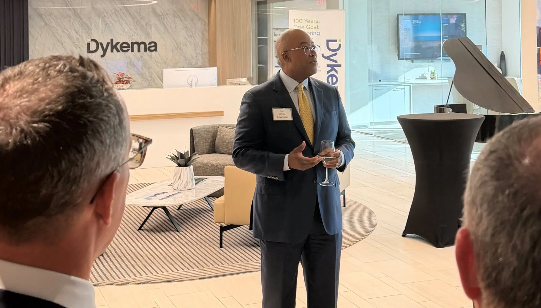A man in a suit and glasses standing in a modern office lobby, holding a glass of wine, speaking to a small group of people. There is a Dykema sign on the wall behind him and a grand piano to his right.