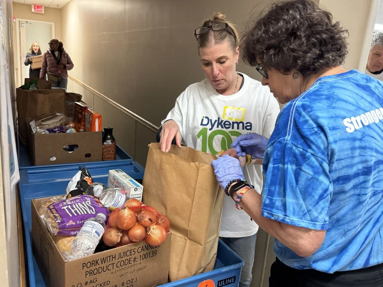 Two women are packing food items into a brown paper bag, with grocery boxes and bottles of water in blue carts nearby. Two other women are in the background holding a box, walking down a hallway.
