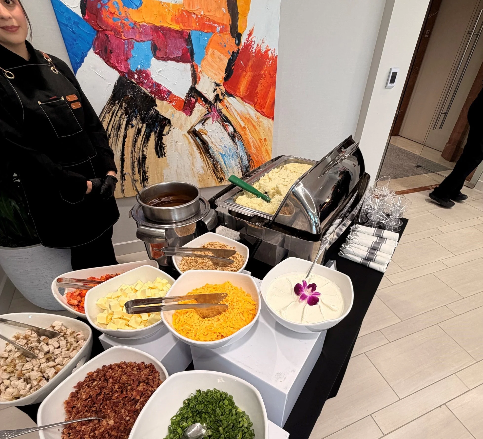 Buffet table with various toppings and ingredients for a dish, including diced cheese, shredded cheese, chopped green onions, and other toppings. In the background, a colorful abstract painting is visible on the wall, and a person dressed in black ch