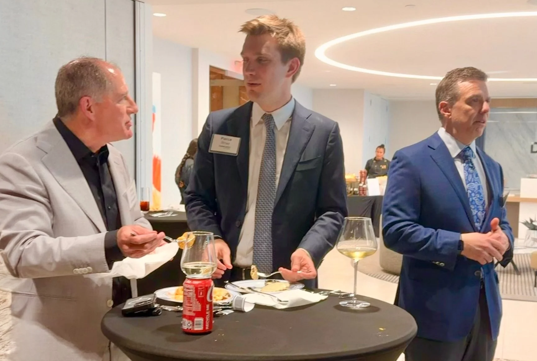 Three men in business attire standing around a tall table with food and drinks, engaged in conversation at a professional event or conference.