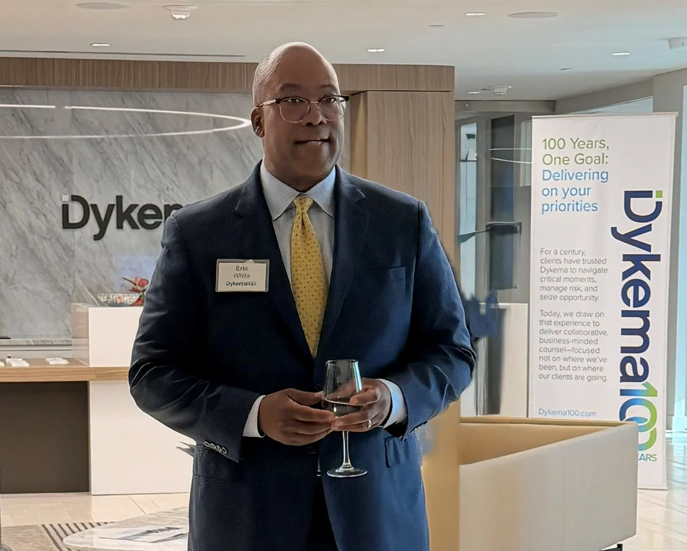 A man in a dark suit and yellow tie holding a glass of water standing in front of a Dykea sign and a reception area, at a professional event or conference.