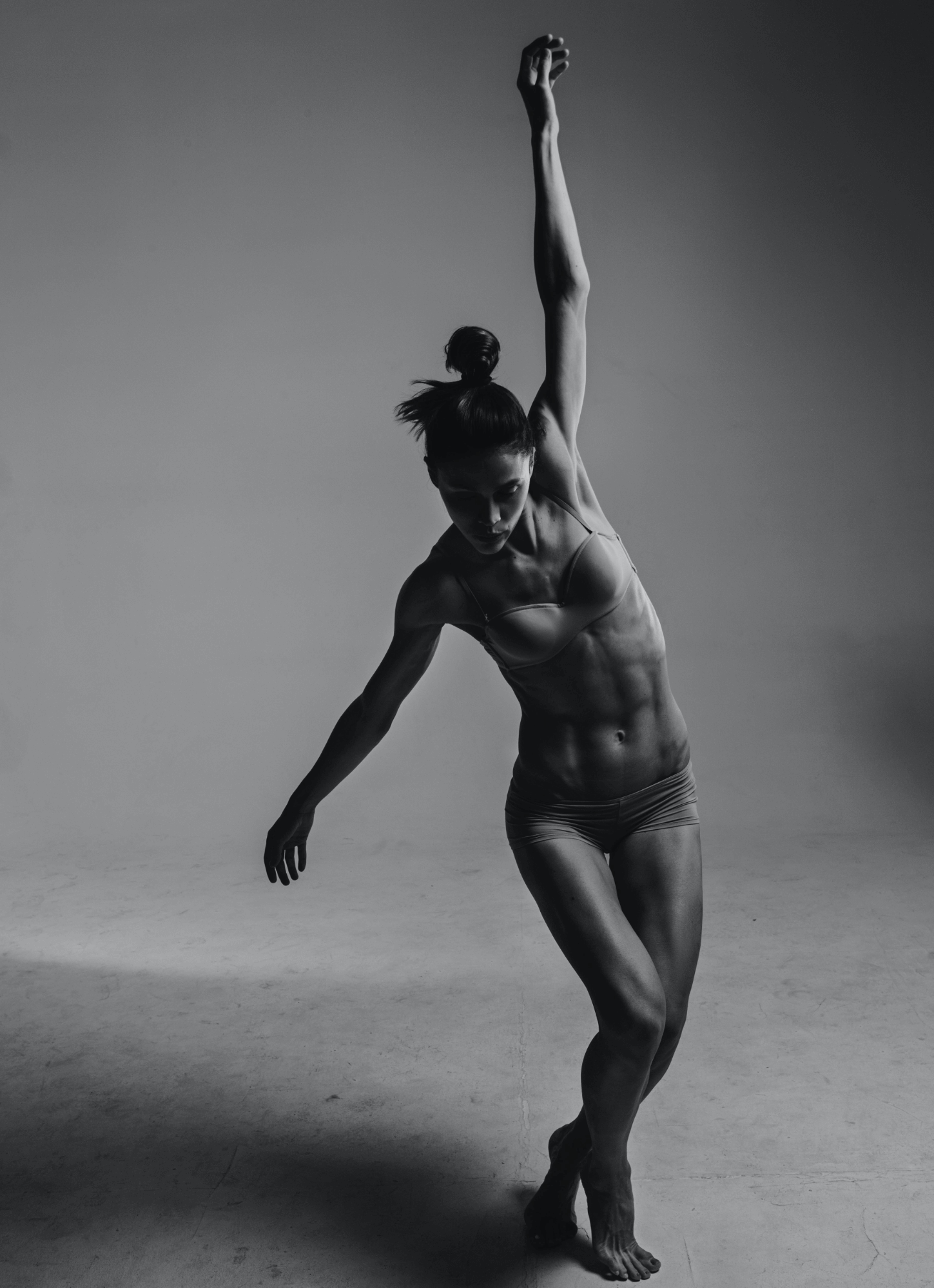 A black-and-white photo of a female dancer in a pose with one arm raised above her head and the other extending downward, wearing a sports bra and shorts, in a studio setting.