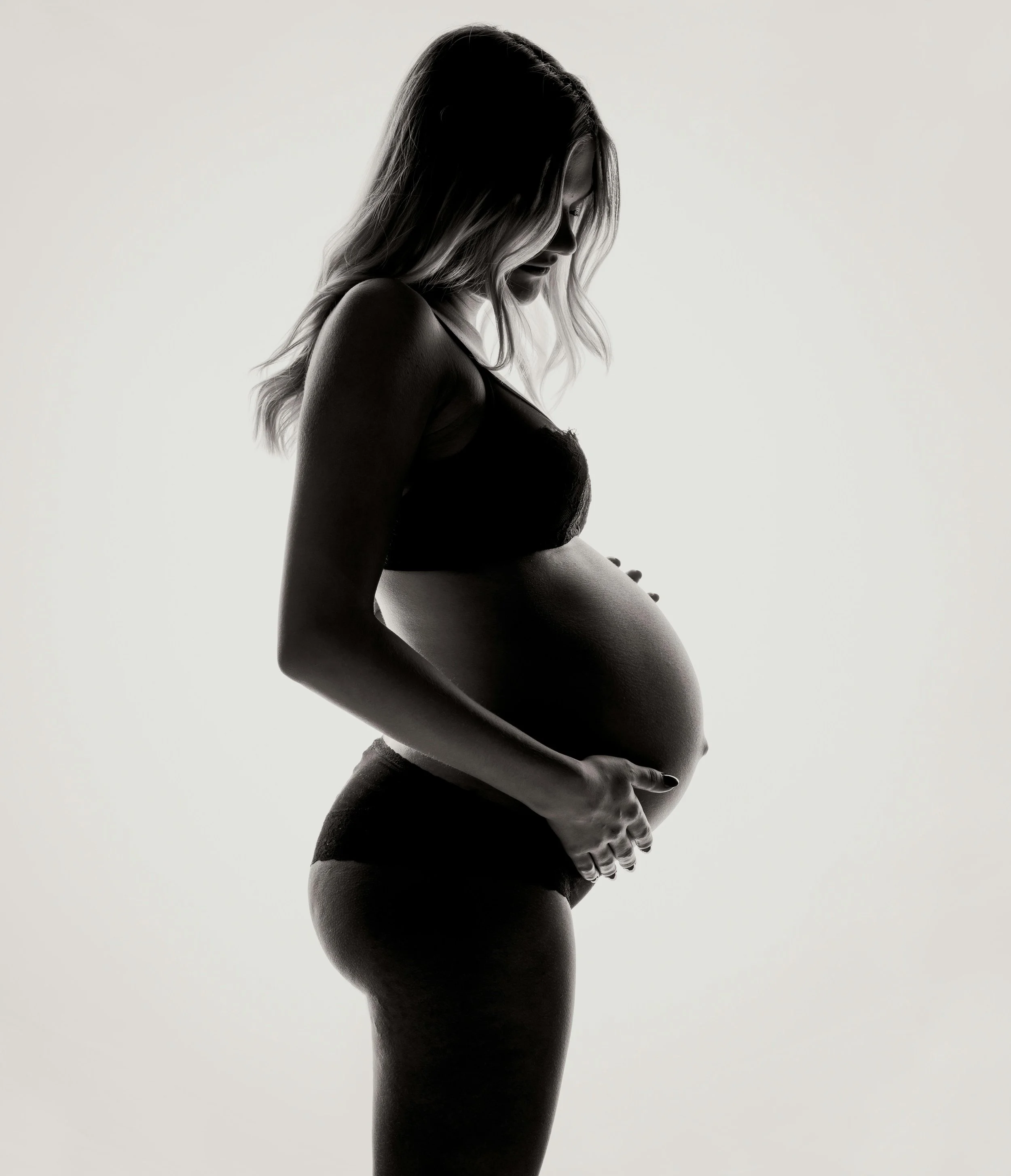 A silhouette of a pregnant woman in black lingerie, gently holding her belly with both hands, standing sideways against a light background.