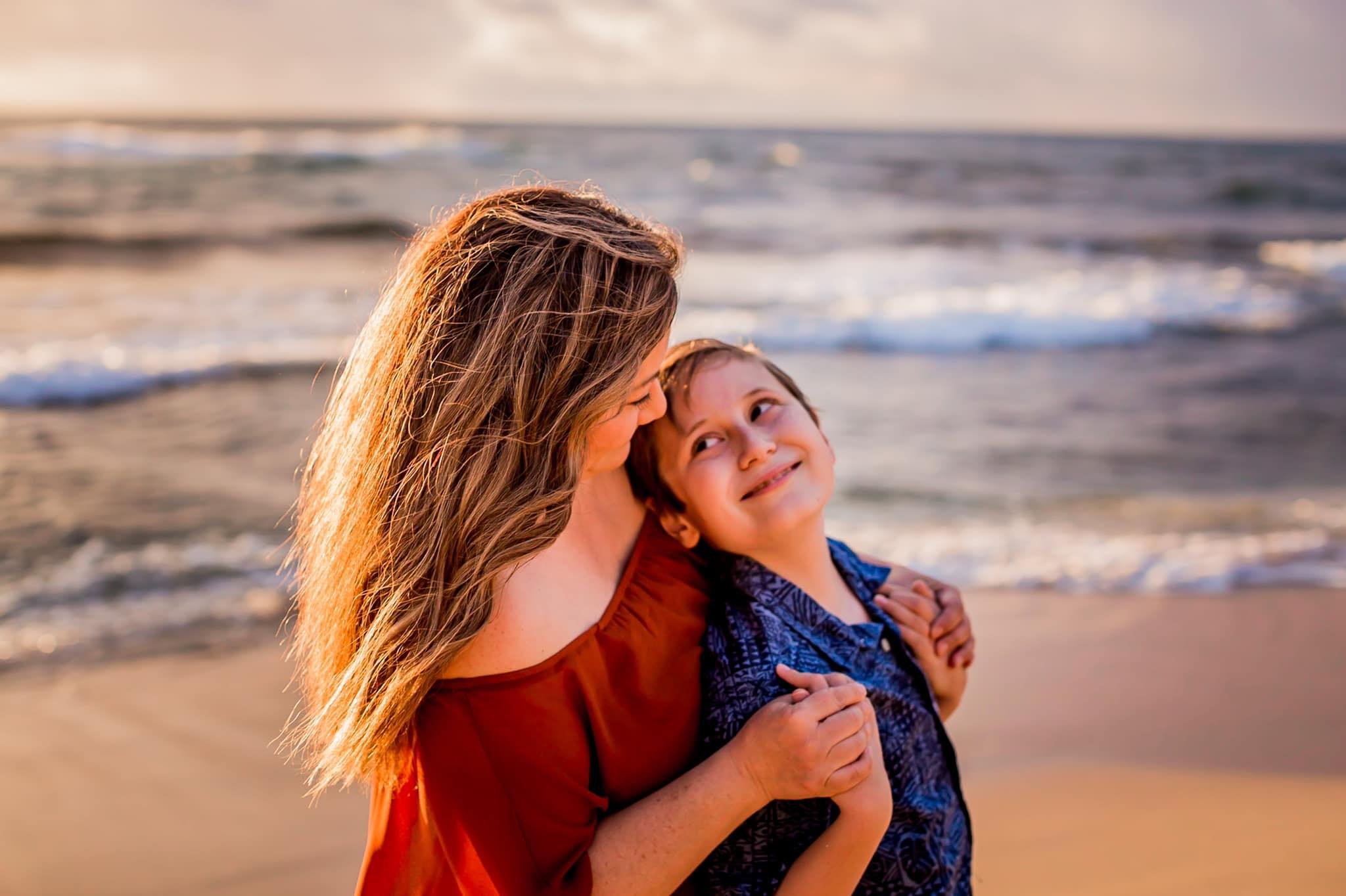 A woman with long brown hair and a young boy with short brown hair at the beach during sunset, smiling and embracing each other.