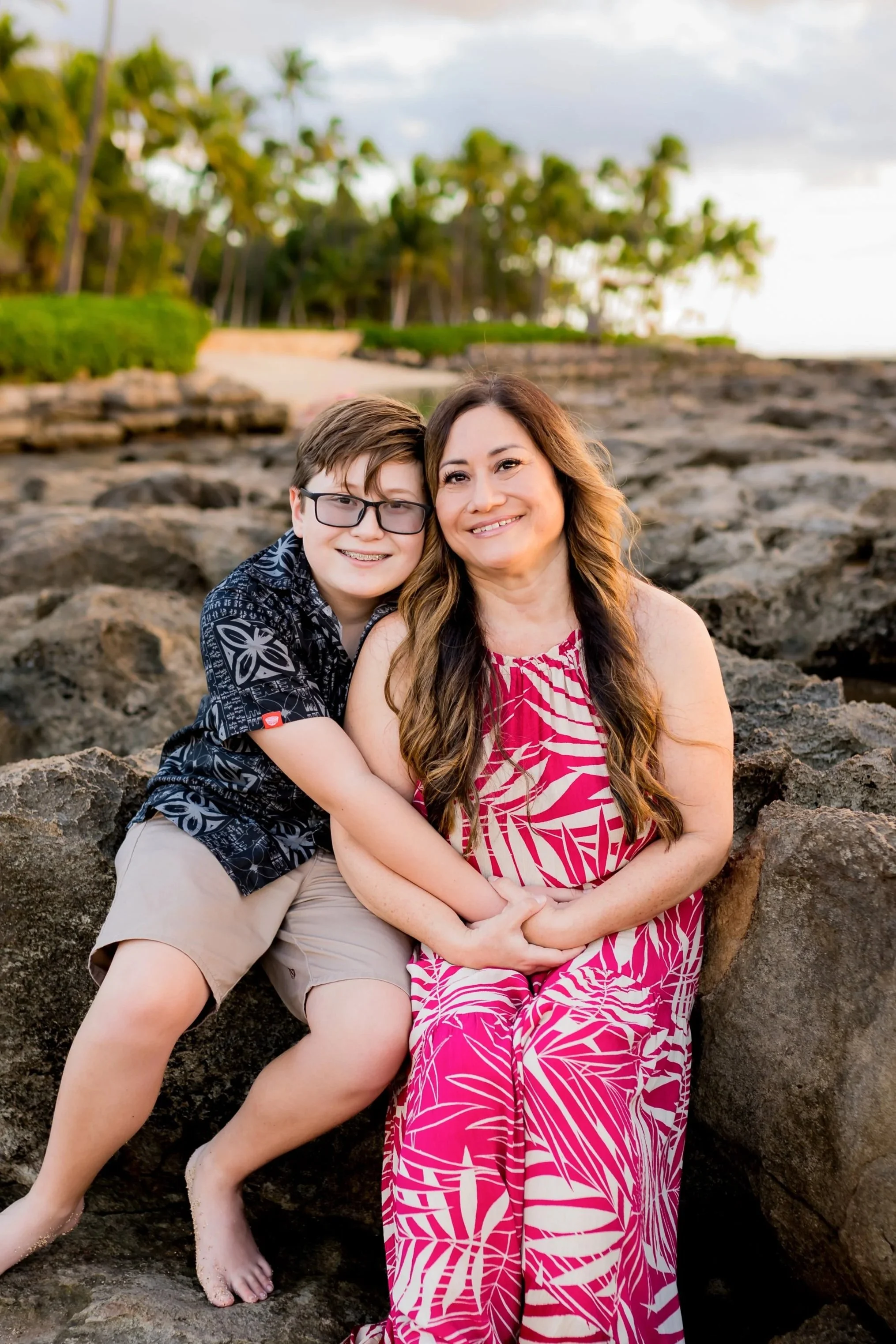 A mother and her son sitting on rocks at the beach, smiling during sunset with palm trees and a grassy area in the background.