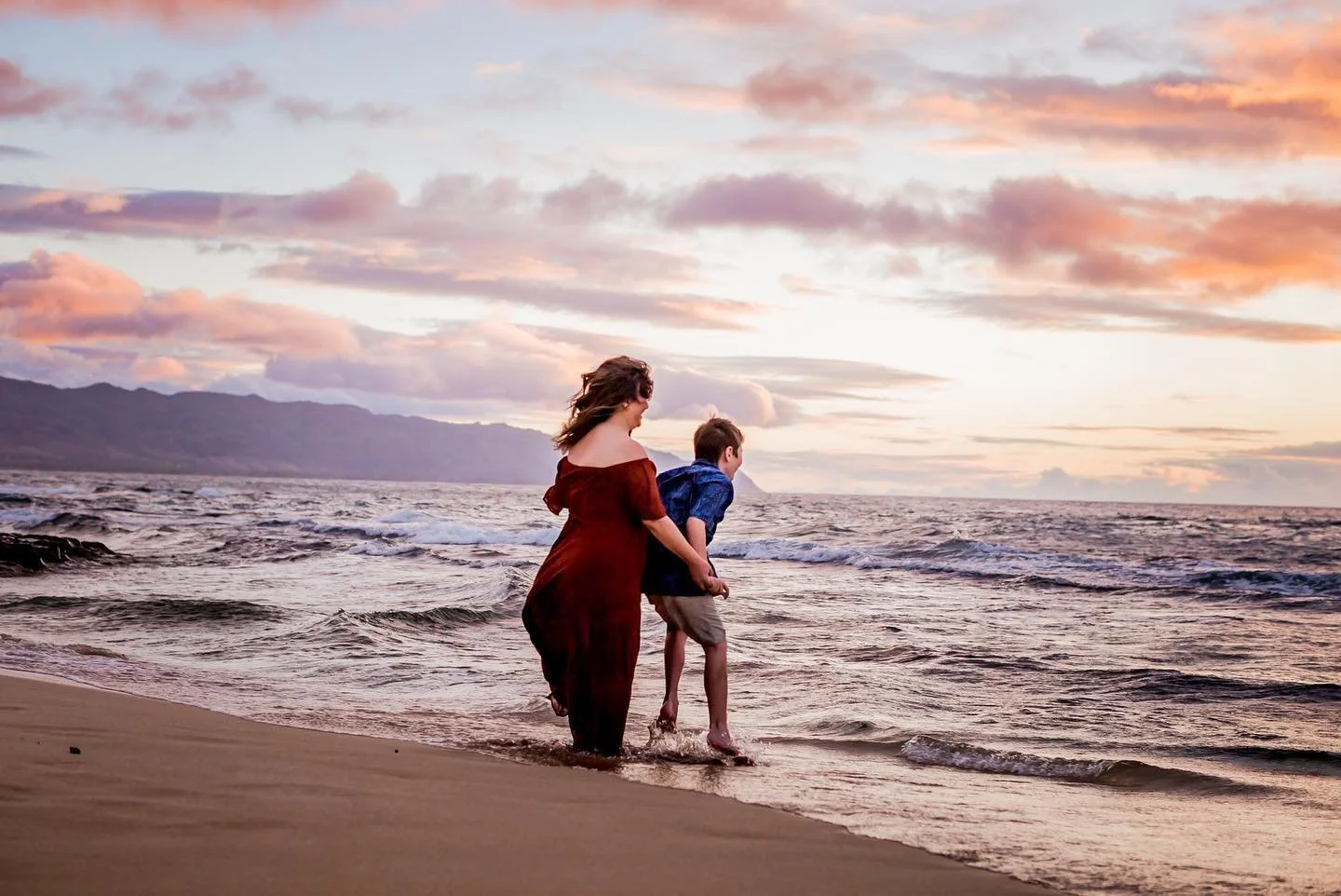 A woman and a young boy walking hand in hand along the beach at sunset, with mountains in the background and waves gently lapping at their feet.