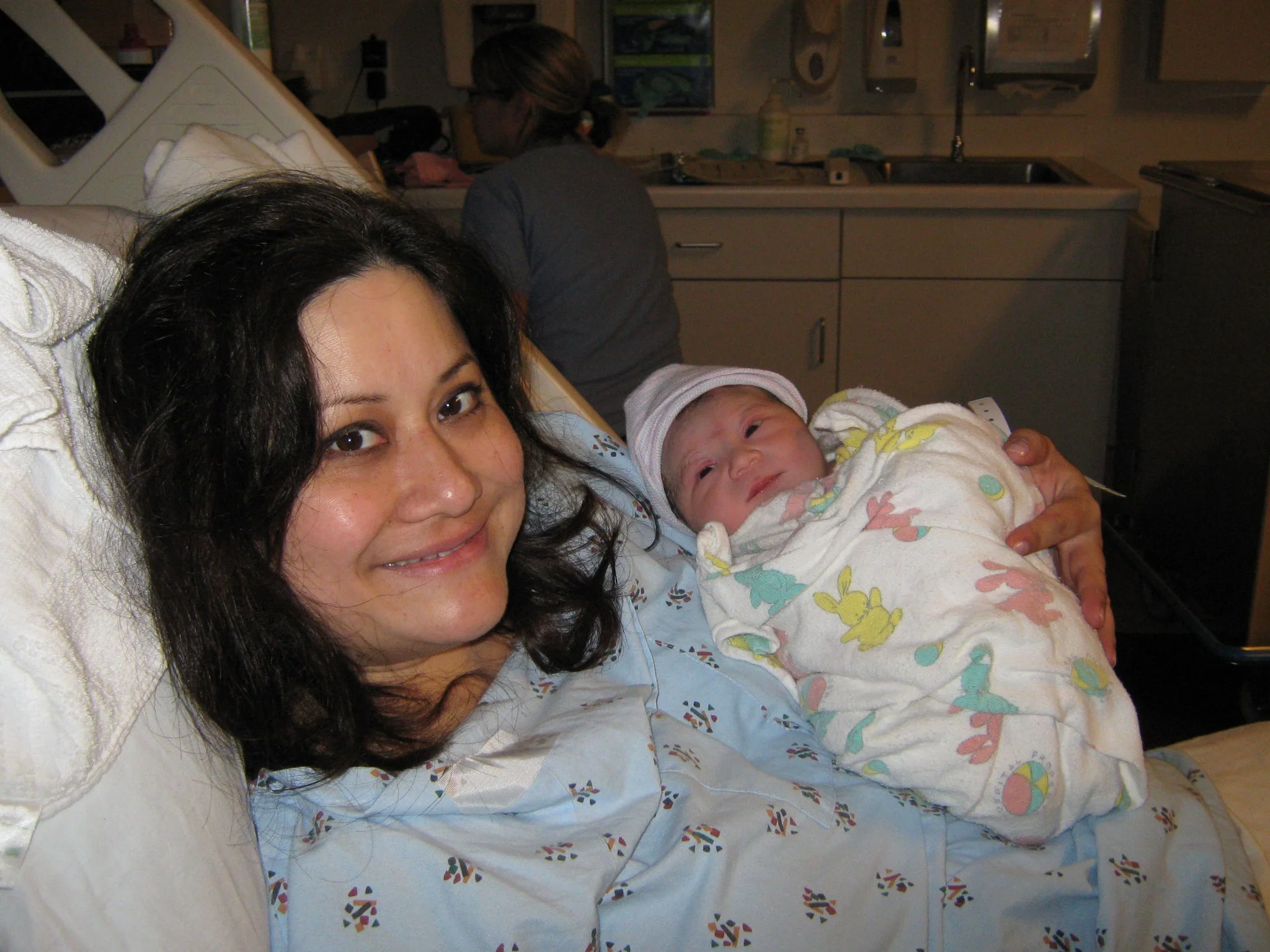 Woman smiling in a hospital bed holding a newborn wrapped in a blanket with pastel bunny and ball patterns, wearing a hospital gown and cap, in a hospital room.