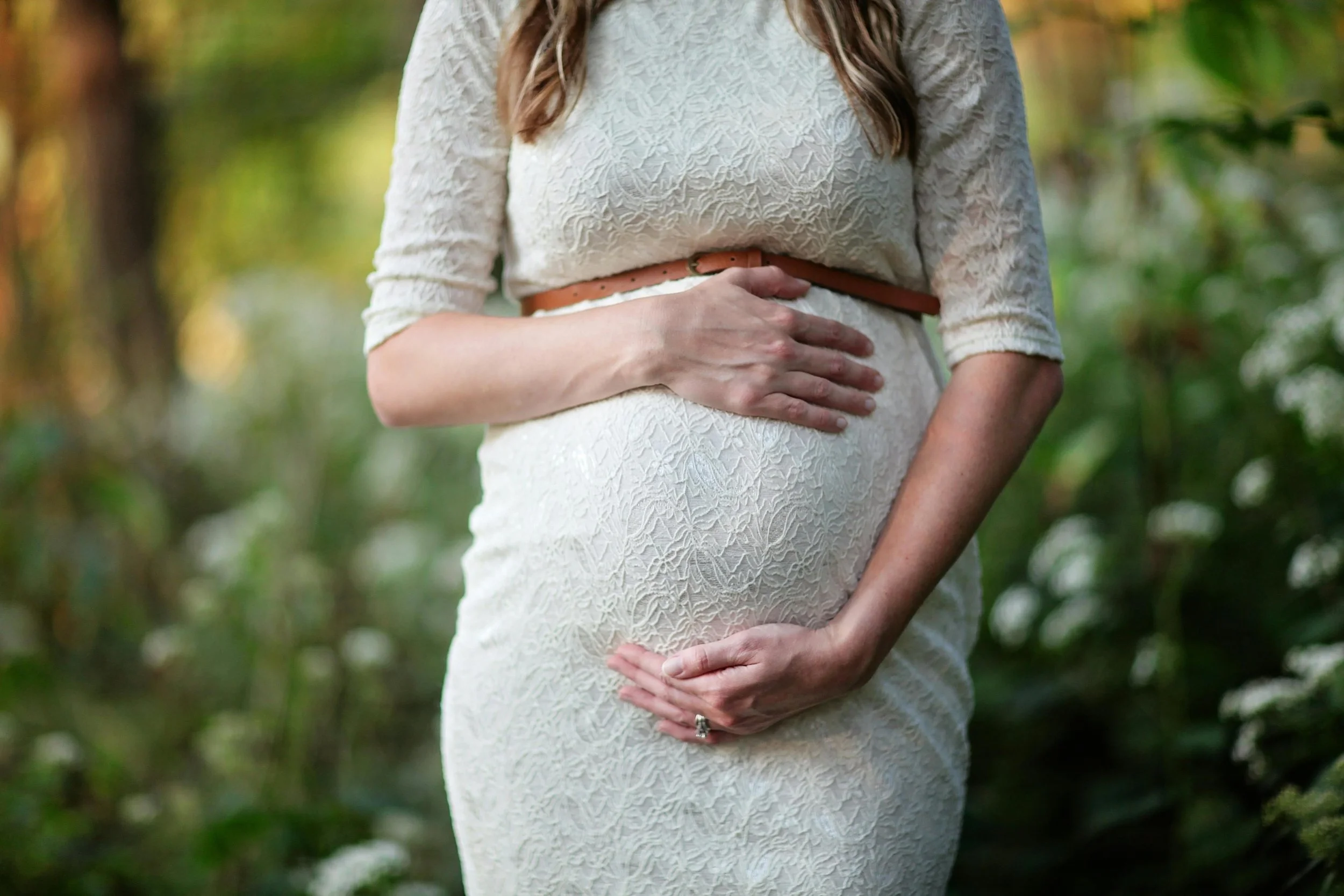 A pregnant woman in a white lace dress outdoors, gently holding her belly with one hand on top and the other below.