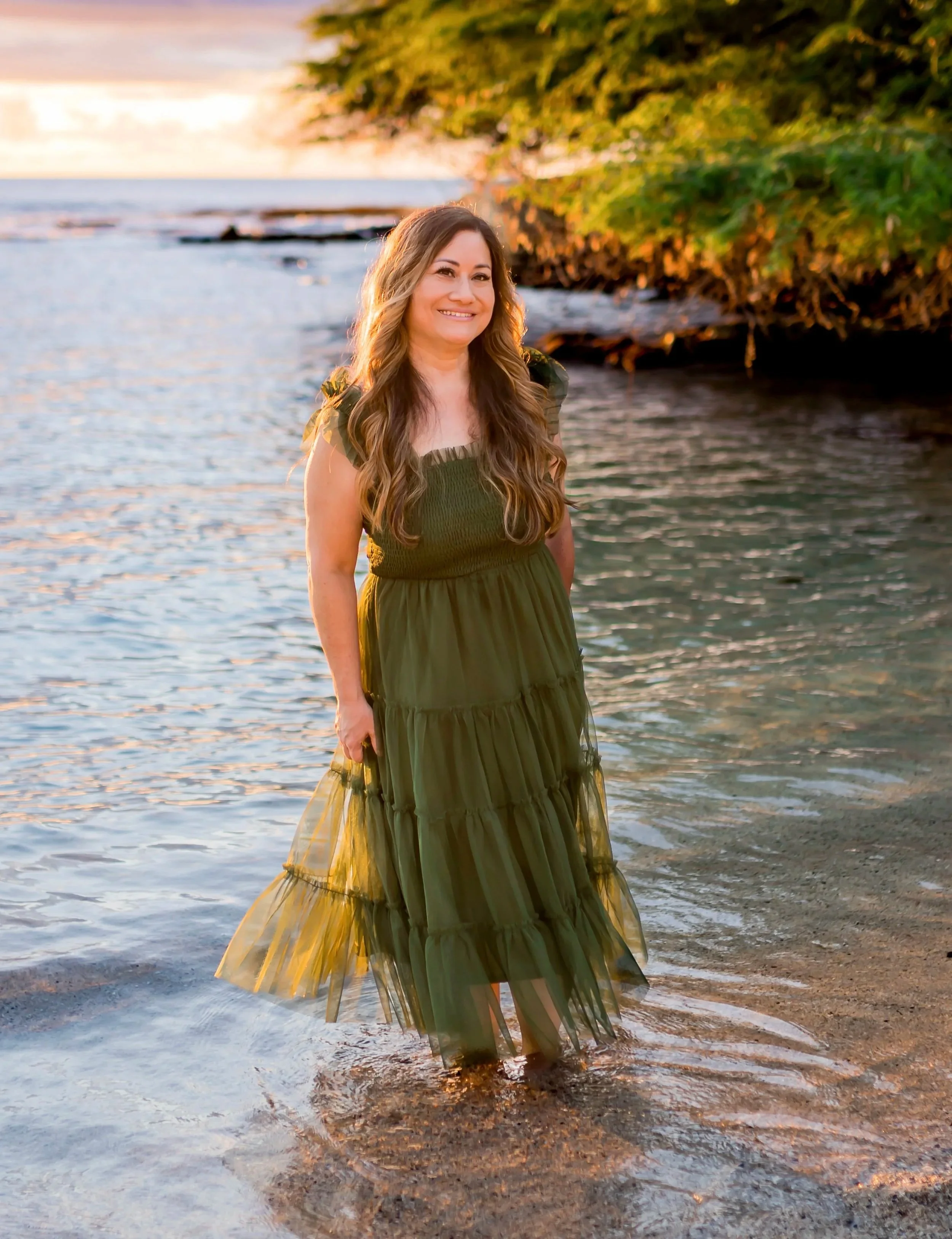Woman in a long green dress standing in shallow water near a tree-lined shoreline during sunset.