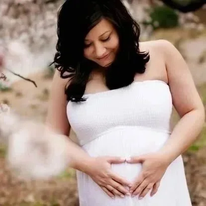 Pregnant woman standing outdoors in a white strapless dress, holding her belly with both hands and looking down.