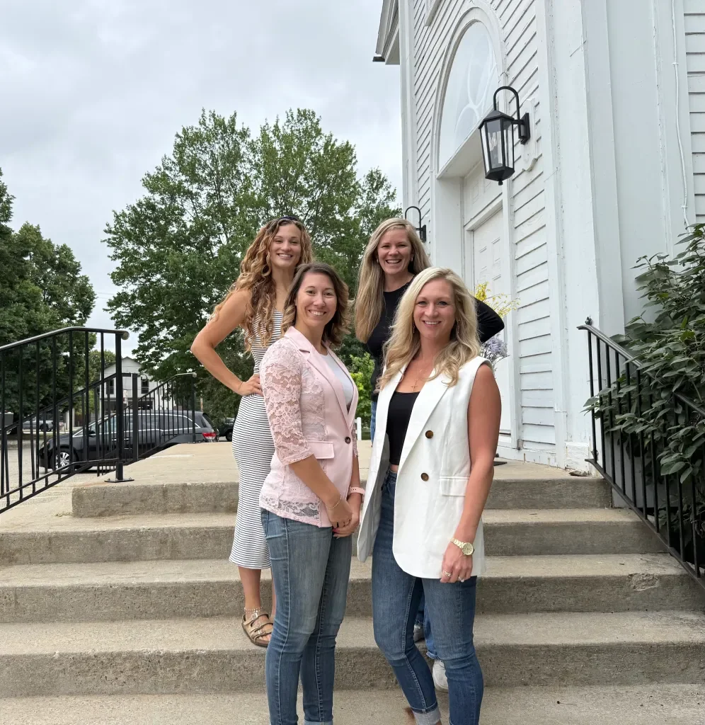 Four women smiling and standing on steps outside a white building with black lanterns, greenery, and parked cars in the background.
